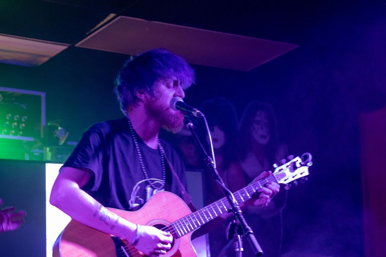 A musician with facial hair plays an acoustic guitar and sings into a microphone under purple stage lighting.