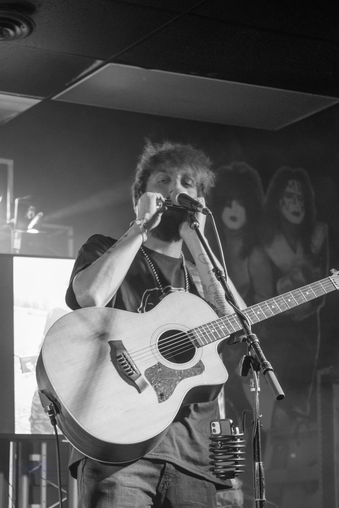 A musician plays an acoustic guitar and harmonica into a microphone on stage in front of a cutout of band members.
