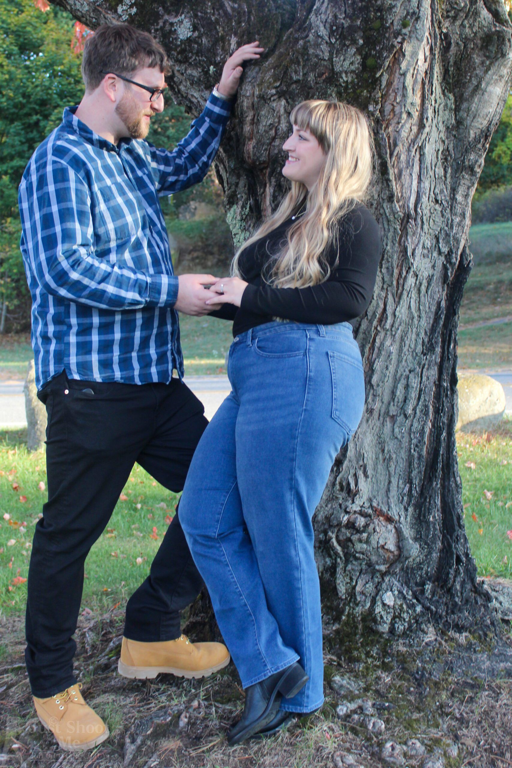 A man in a plaid shirt and a woman in a black shirt stand holding hands by a tree in a park-like setting.