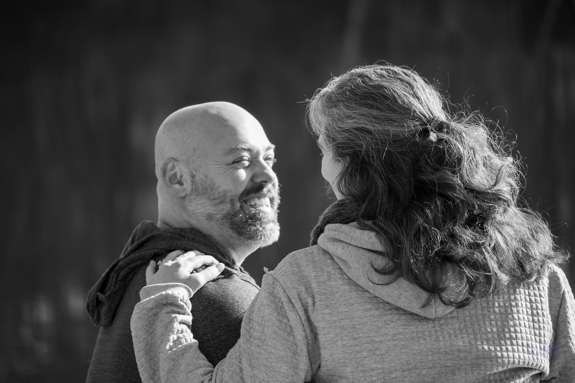A black-and-white portrait of a man smiling at a person who has their arm around him, seen from behind.