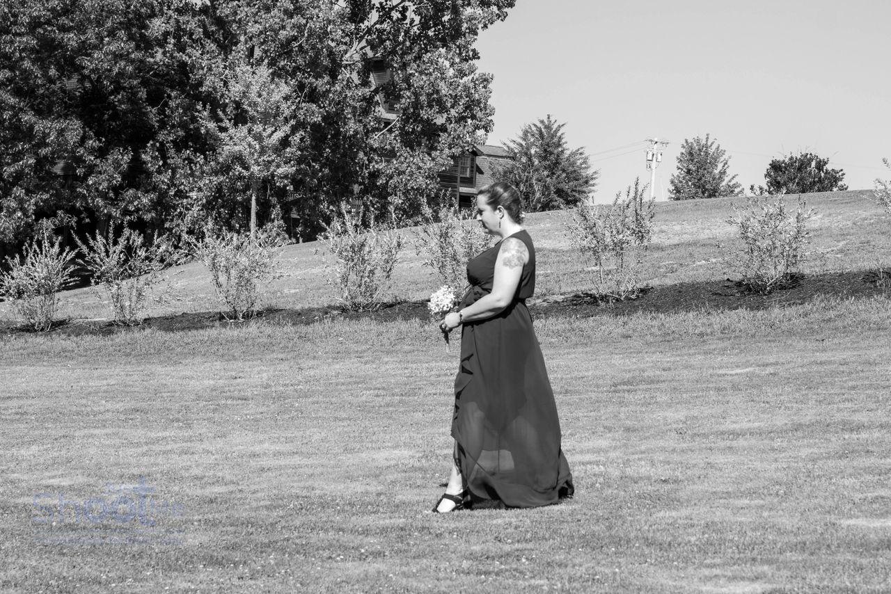 A person in a flowing dress walks across a grassy field with trees in the background.