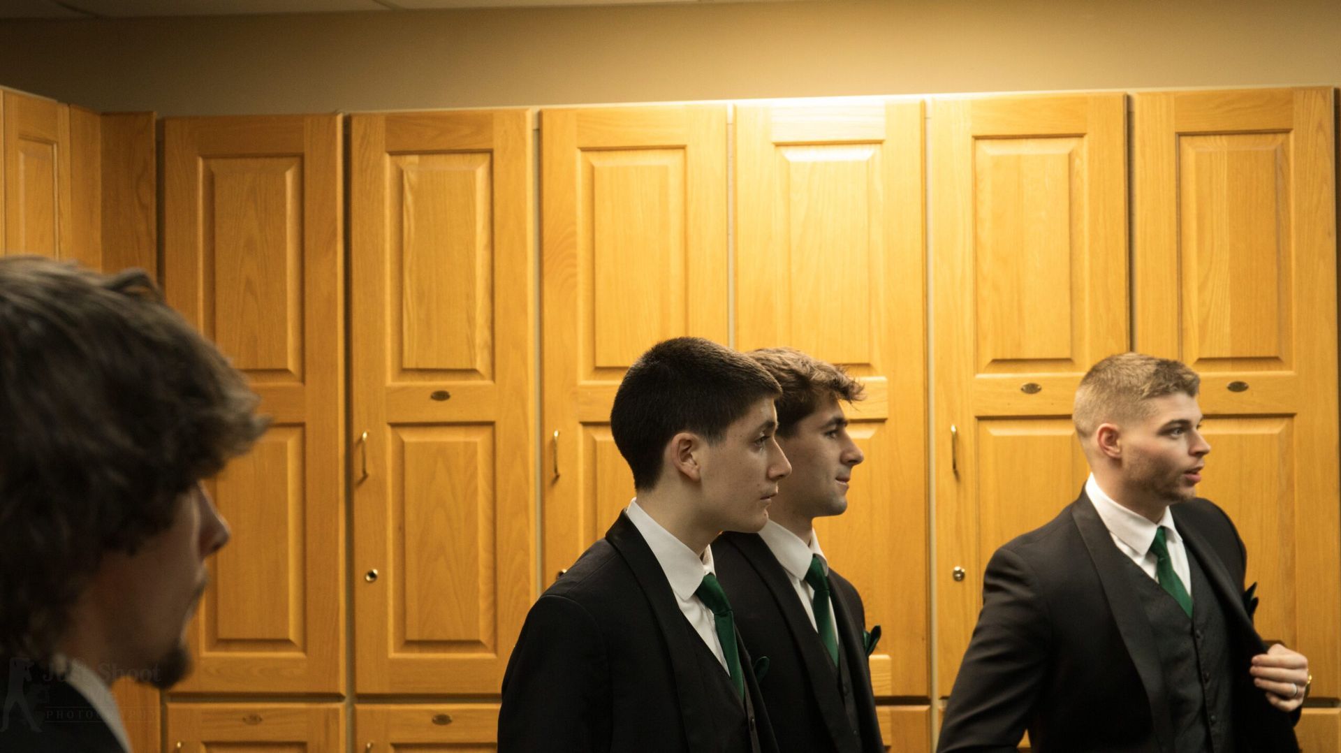 Four individuals in dark suits with green ties stand in front of wooden lockers.