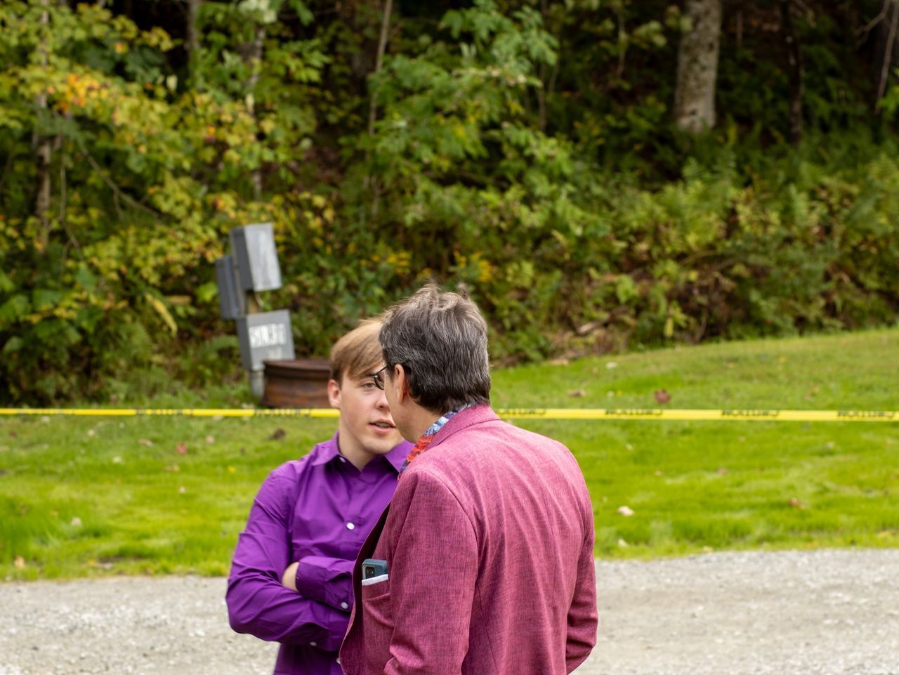 Two people in bright purple and pink shirts stand outdoors, facing each other in conversation near a grassy area.