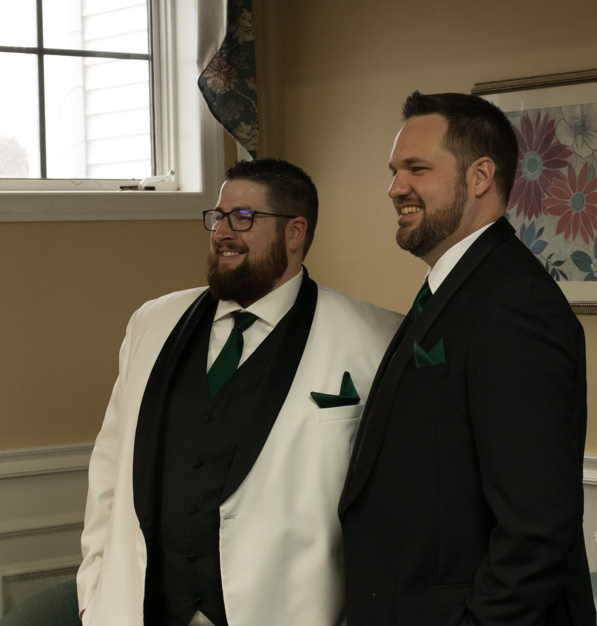 Two people smiling while standing together indoors, one in a white tuxedo with a black vest, the other in a black suit.