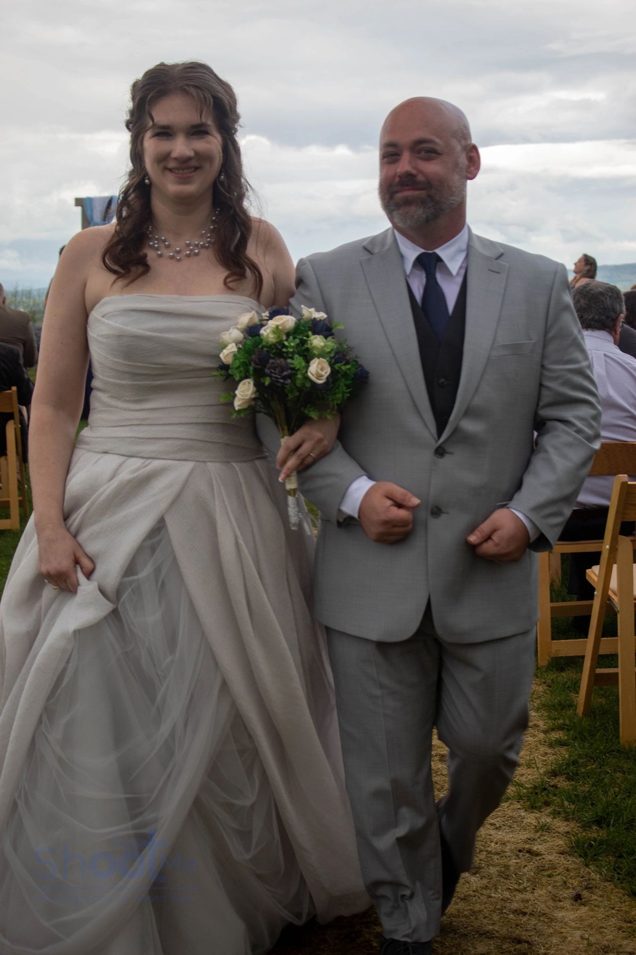 A bride in a strapless gown and a person in a light grey suit walk arm-in-arm outdoors holding a bouquet of flowers.