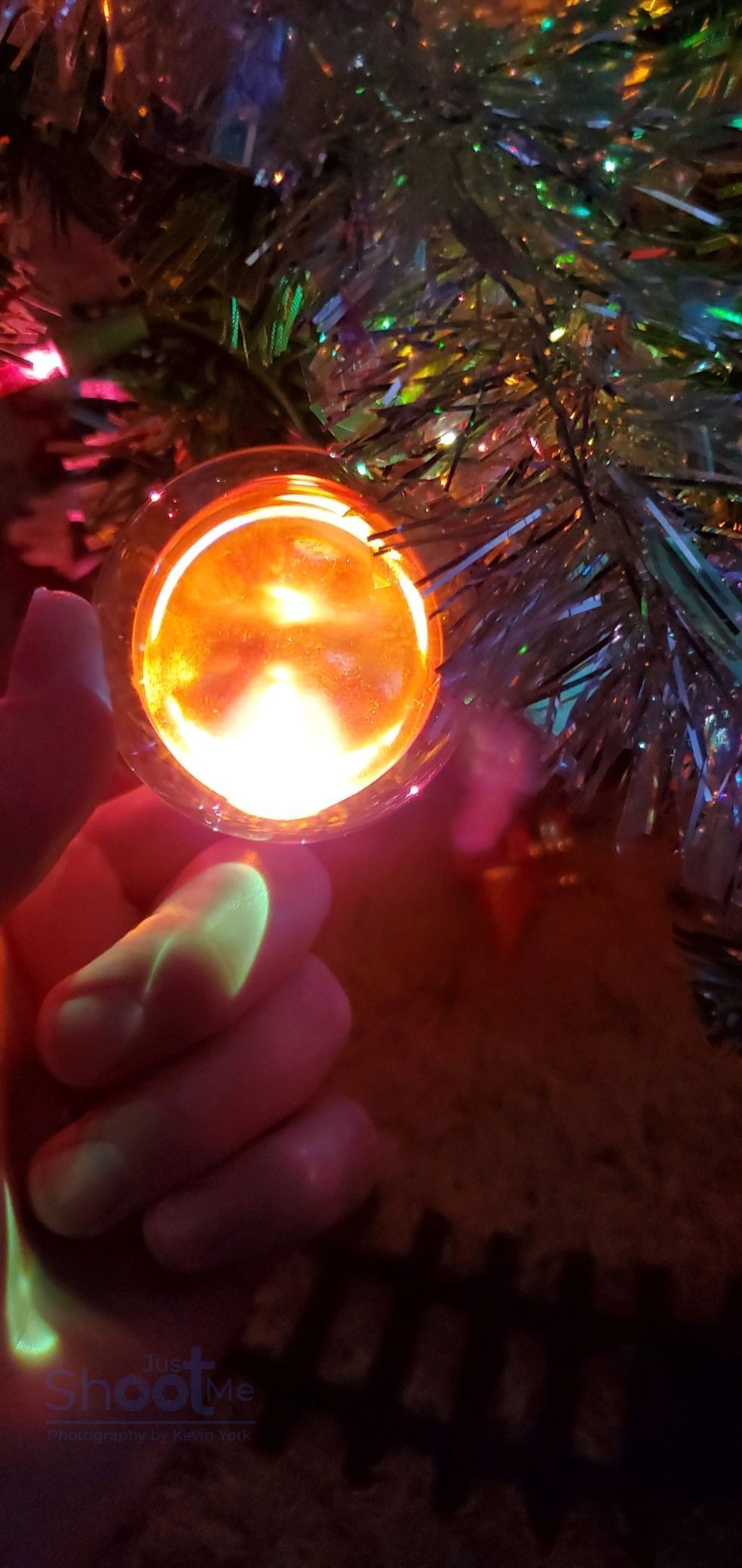 A person holds a bright, circular orange light in front of a Christmas tree with tinsel and model train tracks below.