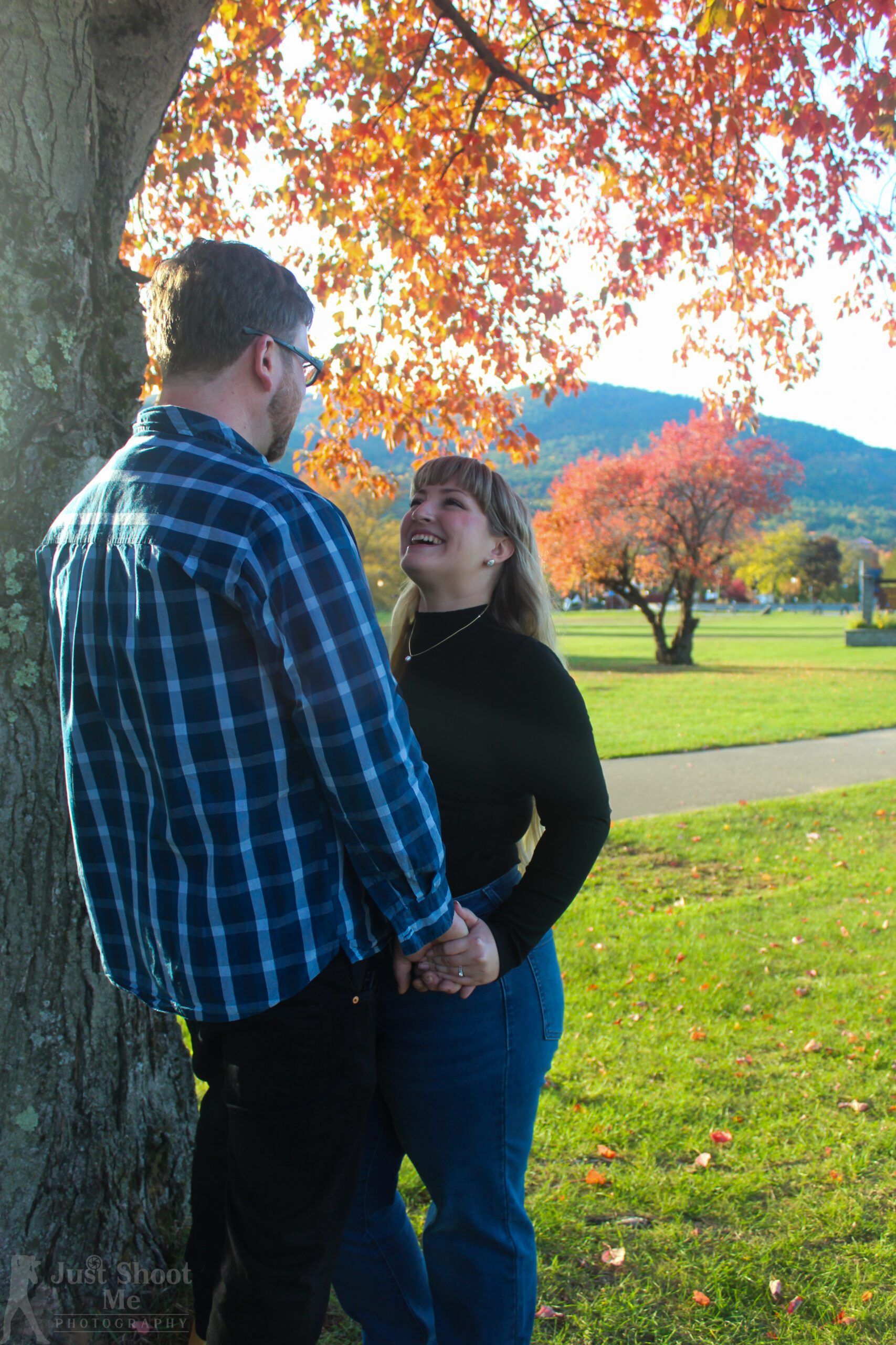 A couple stands holding hands by a tree during autumn, looking at each other beneath vibrant orange-leaved branches.