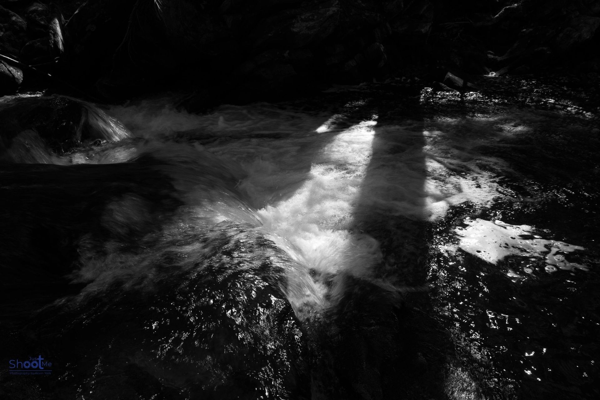 A black and white close-up of turbulent water rushing over dark rocks, with a sharp diagonal shadow cast across the center.