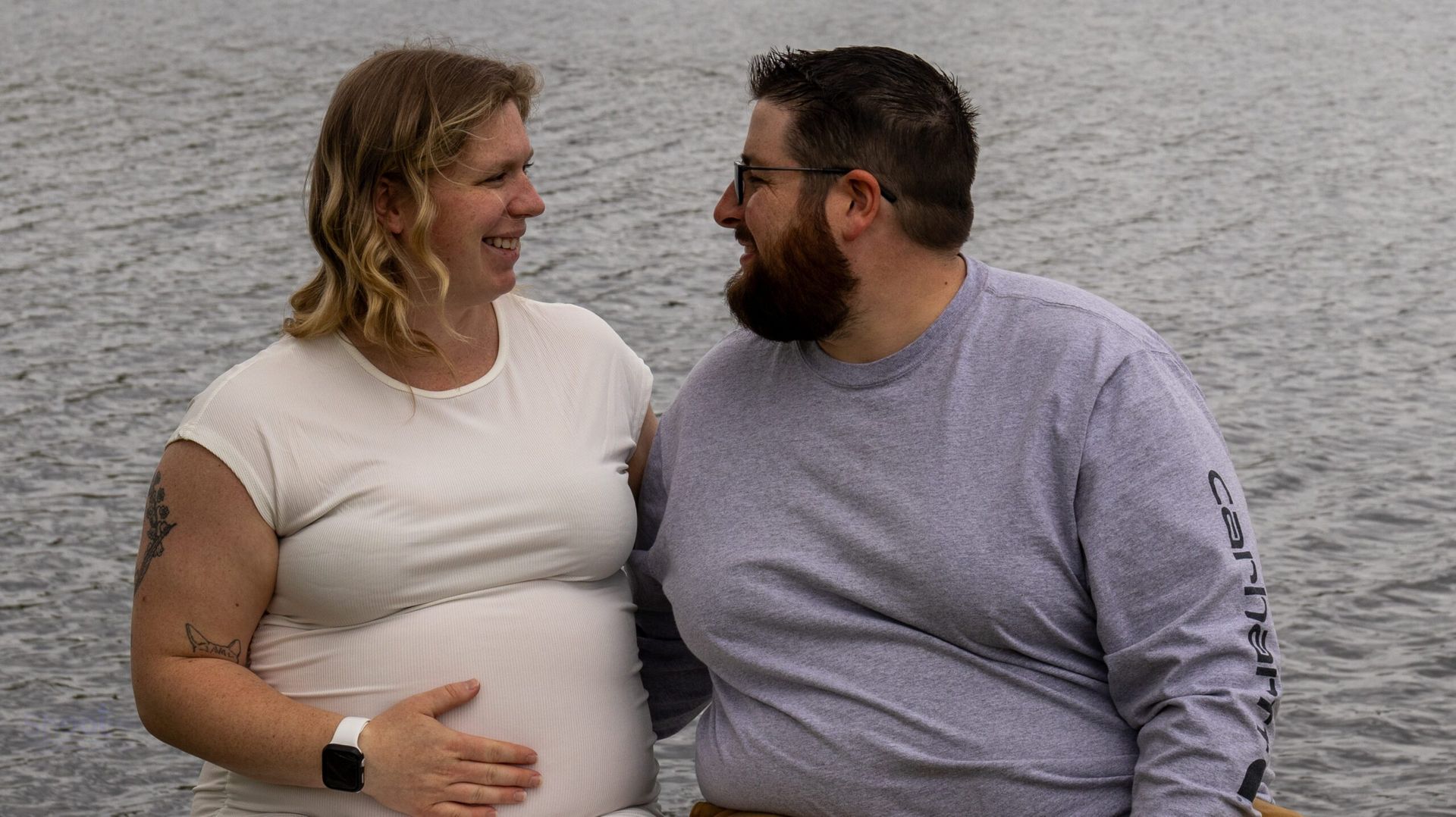 A person with a baby bump and a person with a beard smiling at each other by the water.
