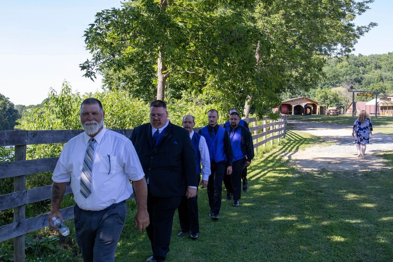 A line of people in formal wedding attire walking along a rural wooden fence on a sunny, grassy path.