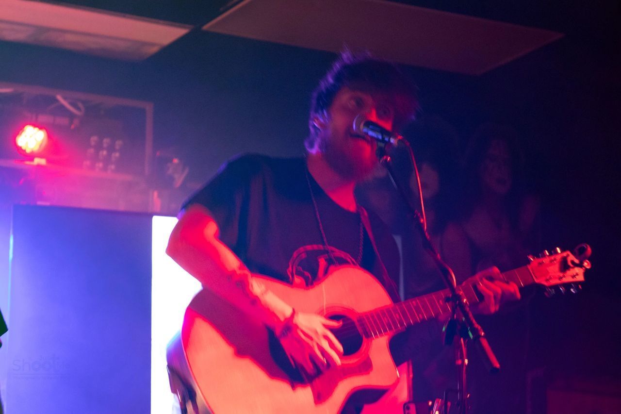A performer sings into a microphone while playing an acoustic guitar on a stage lit with red and blue light.