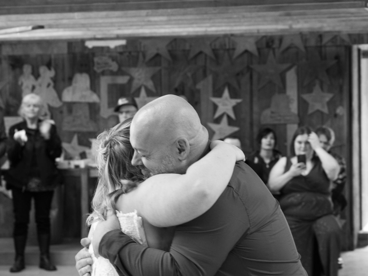 A person in a dark shirt embraces someone in a white garment, with blurred guests watching in the background.
