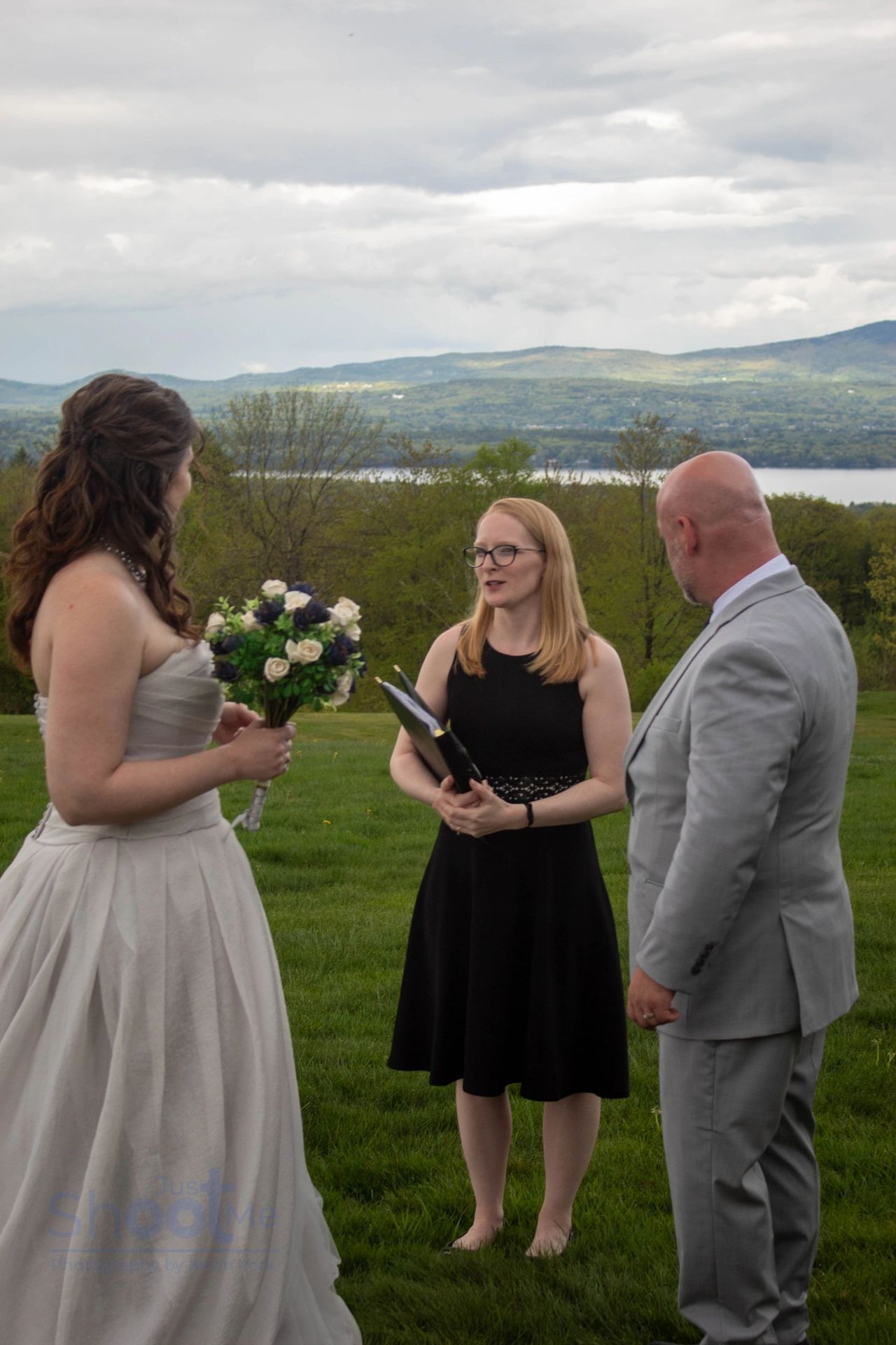 A wedding ceremony officiant stands between a bride in a white dress and a groom in a gray suit overlooking a landscape.