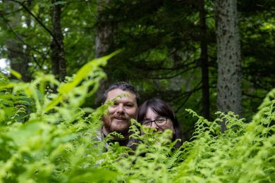 Two people looking toward the camera from behind a large patch of lush green ferns in a wooded forest setting.