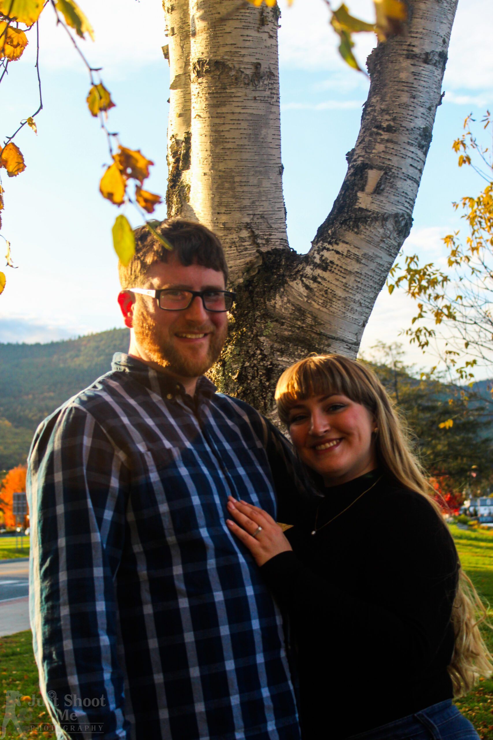 A couple standing close together smiling in front of a white birch tree in an outdoor autumn setting.