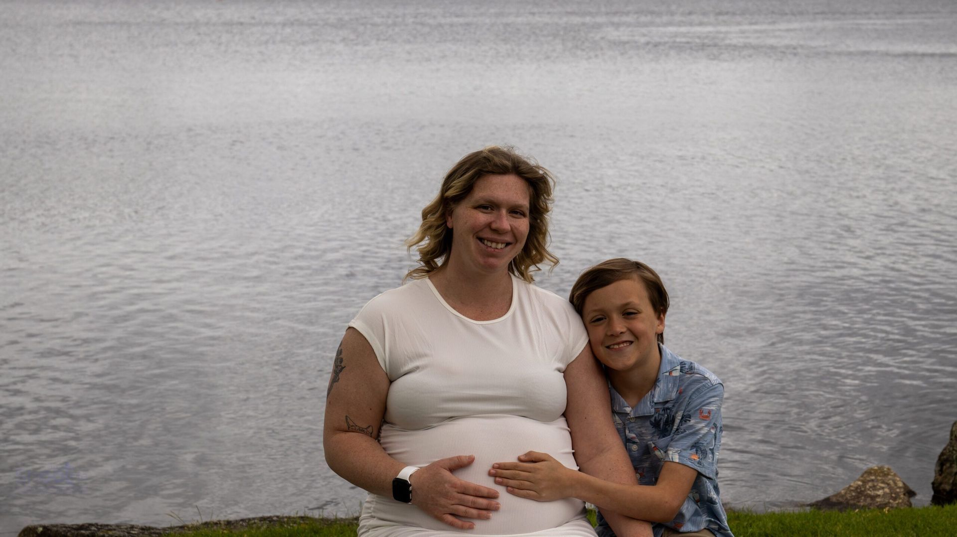A person with light brown hair and a child sit together by a lake, resting their hands on the person's pregnant belly.