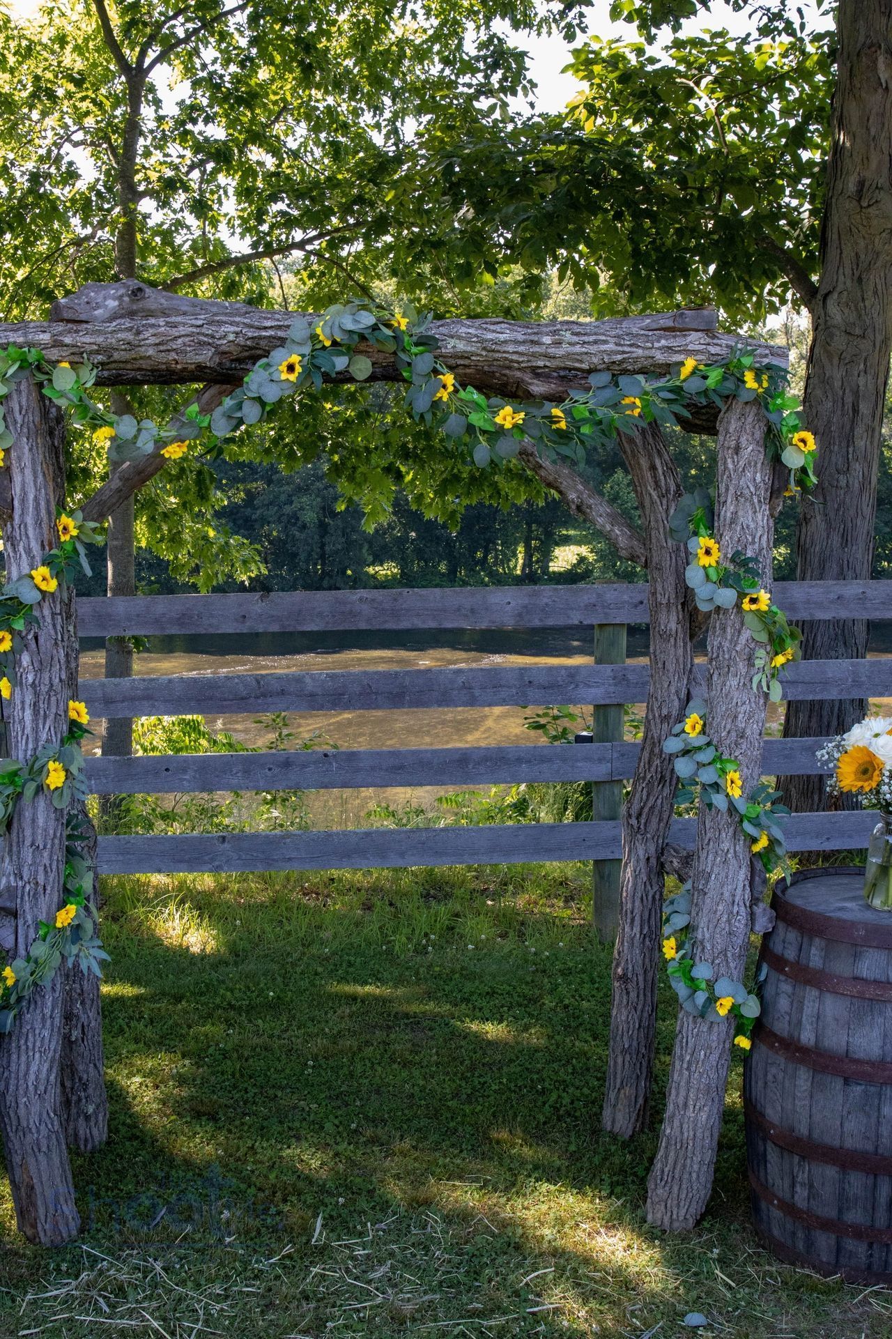 A rustic wooden wedding arch decorated with greenery and sunflowers, standing in a grassy field with a wooden barrel.