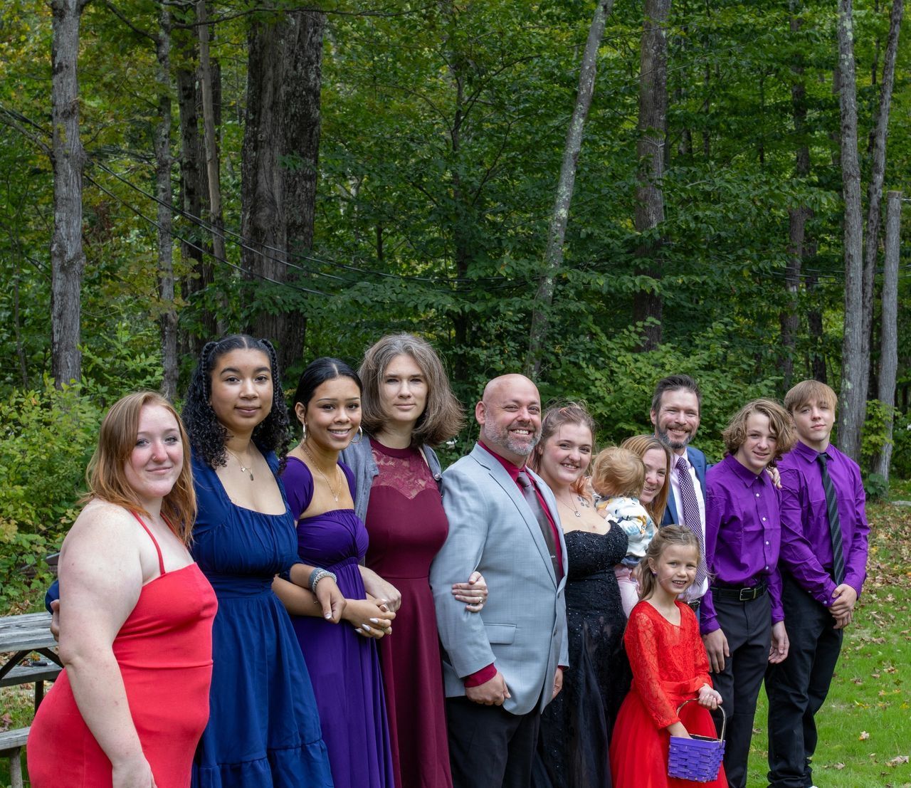 A family standing in a line outdoors against a backdrop of trees, dressed in formal attire in shades of red, blue, and purple.