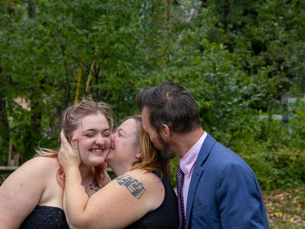 Two people embrace and kiss a smiling person in a black dress outdoors against a green, wooded background.