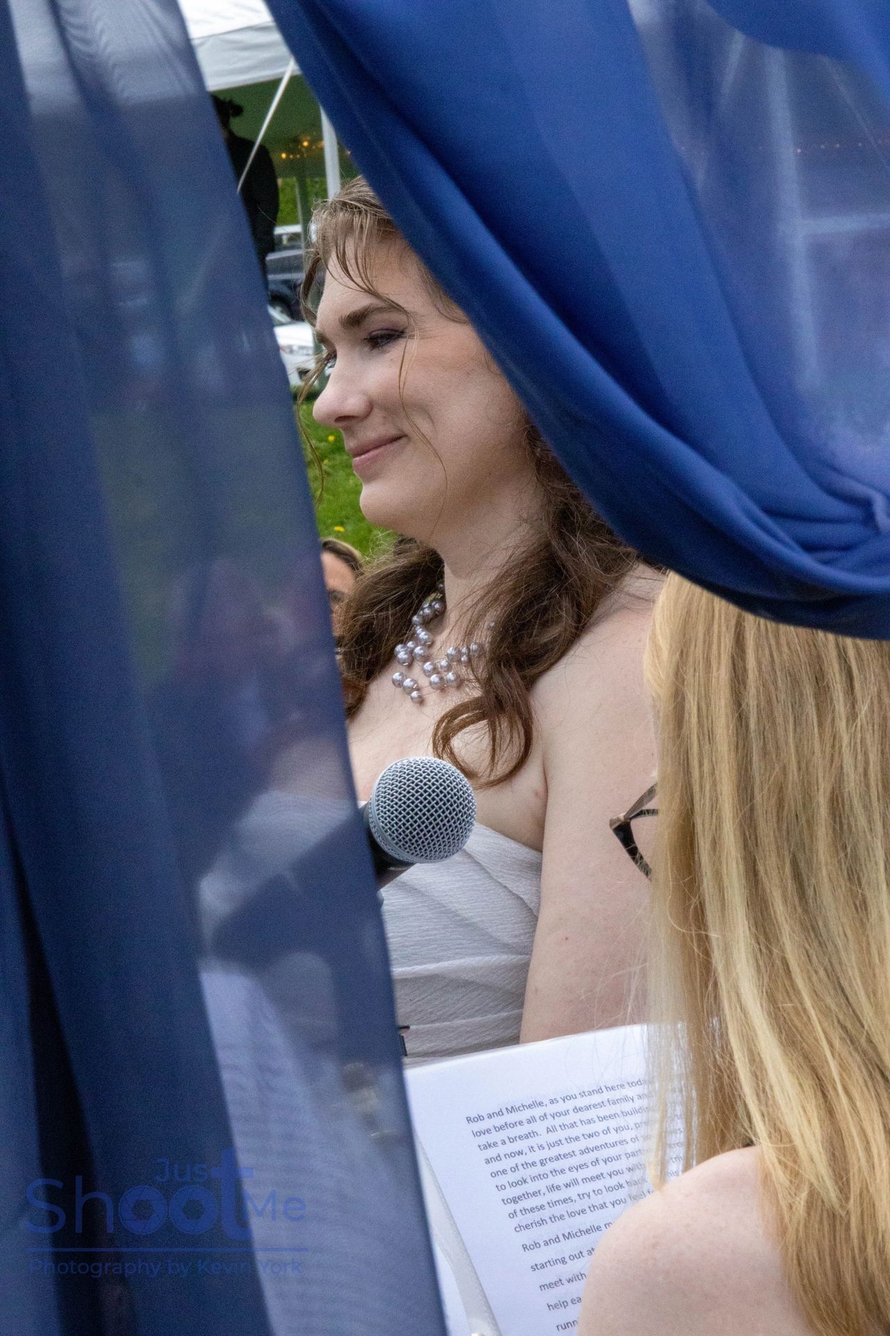 A person smiles while speaking into a microphone at an outdoor wedding, partially framed by blue drapes.