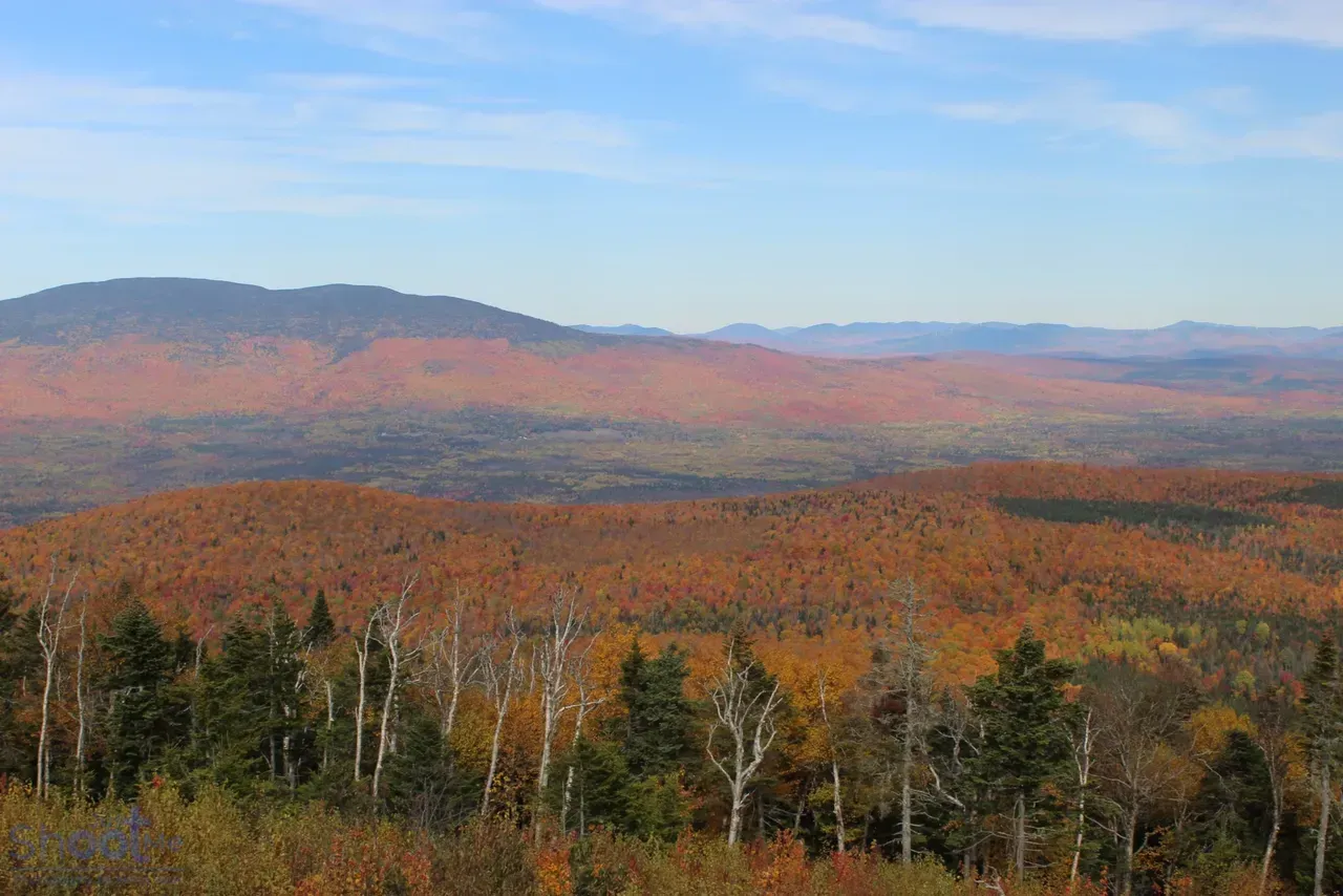 A scenic mountain view with colorful fall foliage, stretching from a foreground of trees to distant, hazy mountain peaks.