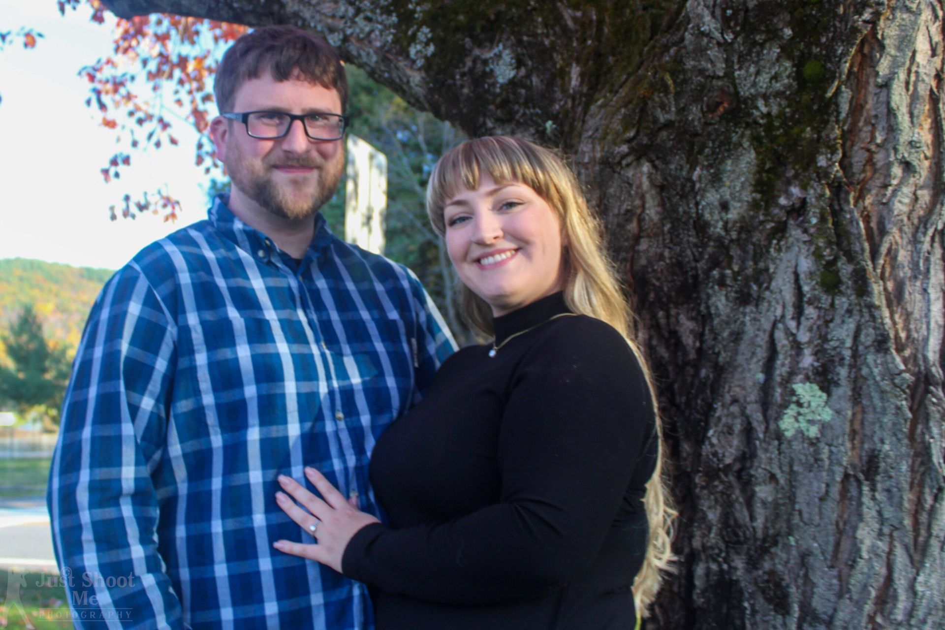 A man in a blue plaid shirt and a woman in a black sweater stand smiling side-by-side in front of a large tree.