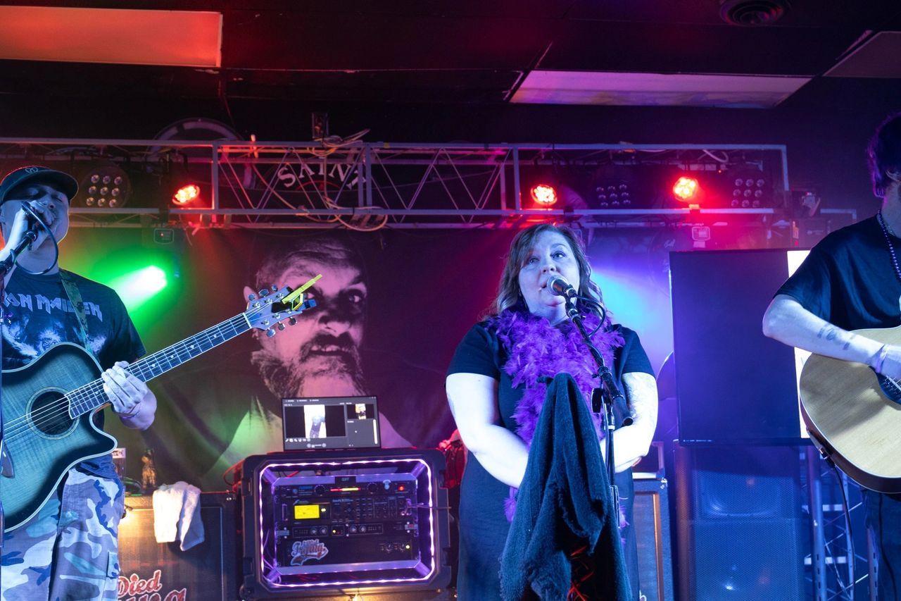 Three musicians perform on a stage with colorful lighting, playing acoustic guitars and singing into microphones.