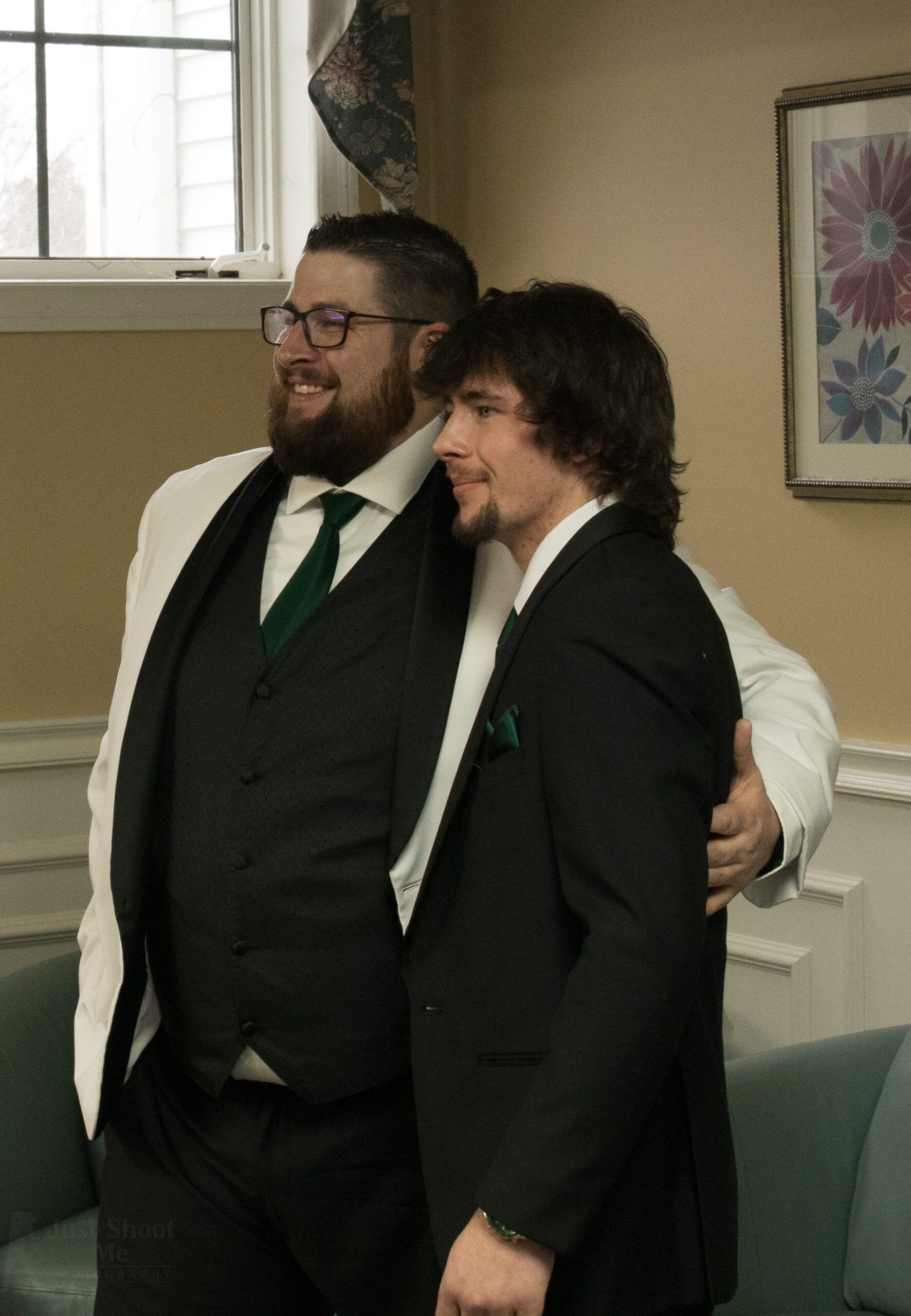 Two men in suits embrace, smiling, inside a room with floral art on the wall.