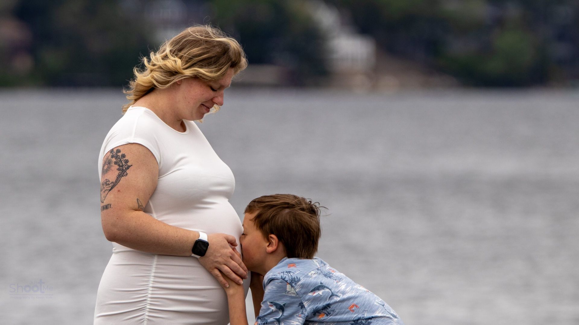 A person in a white top smiles while a child kisses their baby bump by the water.
