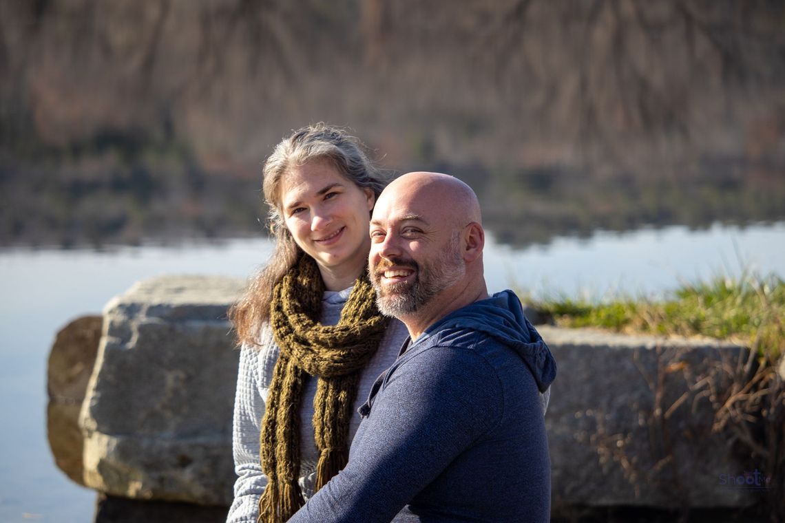 A person with long hair and a scarf poses smiling next to a bald person in a blue hoodie by a lake.