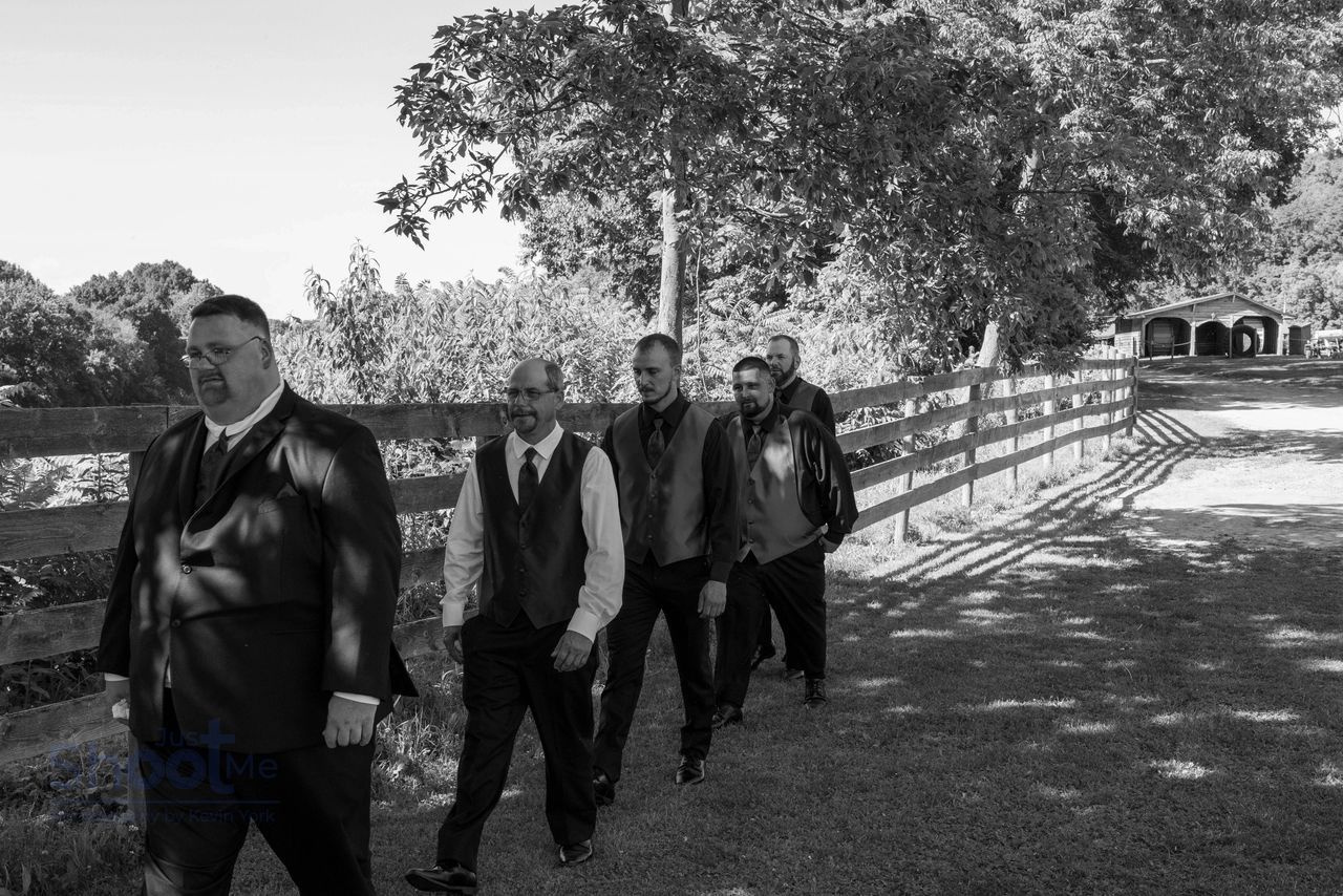 Five men in formal attire walk in a line along a wooden fence on a sunny, outdoor path.
