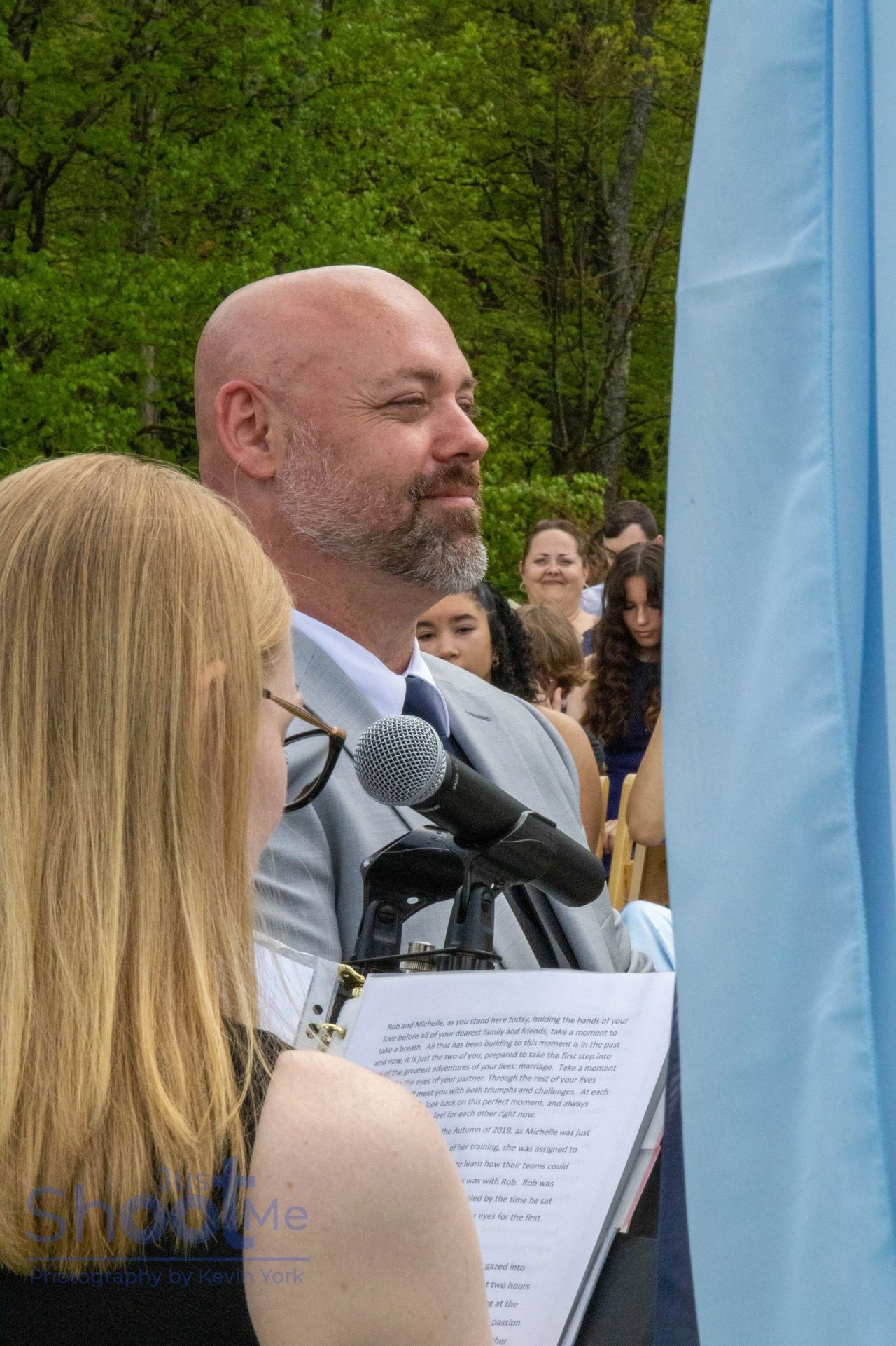 A person in a light suit speaks into a microphone at an outdoor event, with an audience visible in the background.