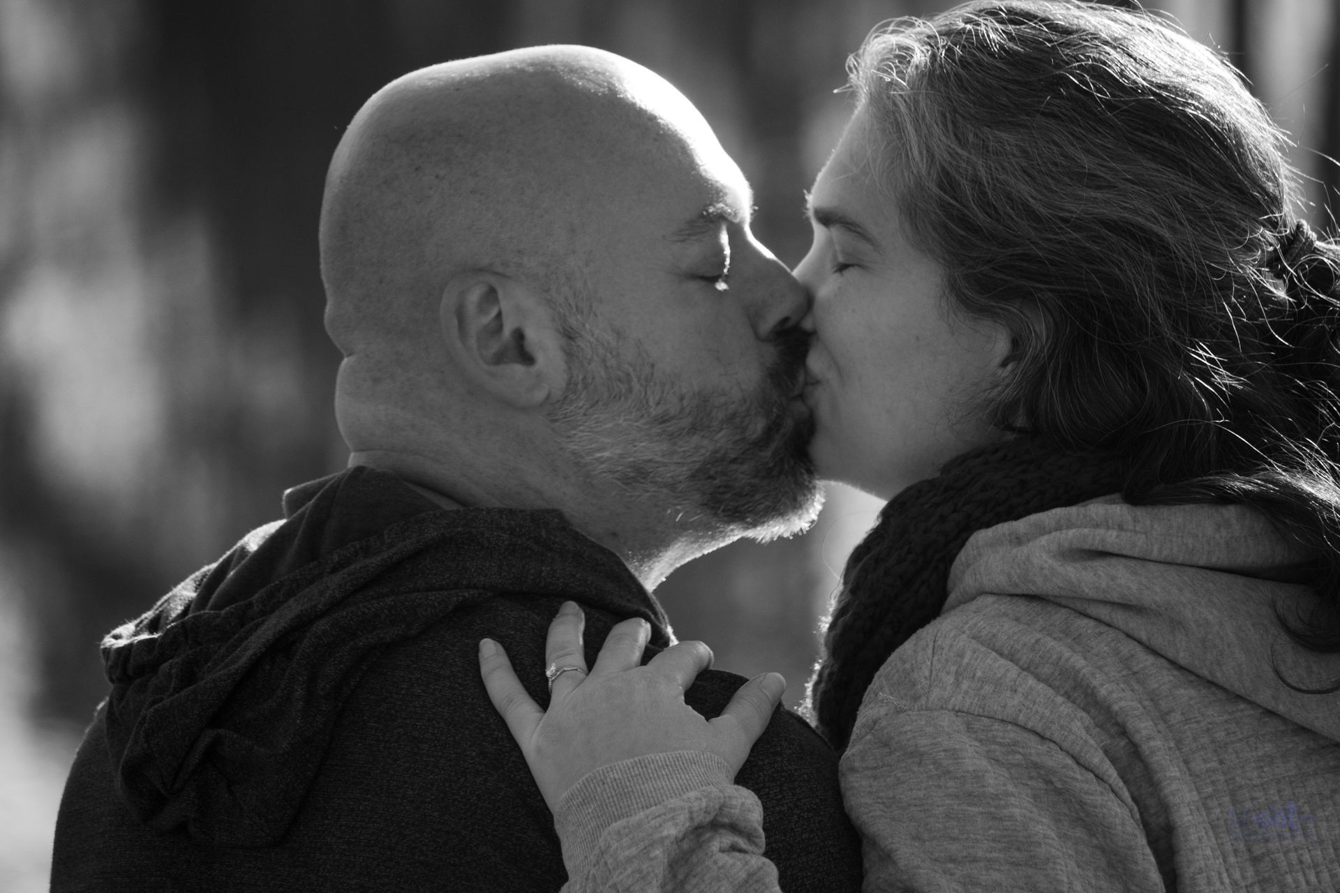 A man and woman sharing a close kiss in profile, both wearing hooded tops, captured in black and white.