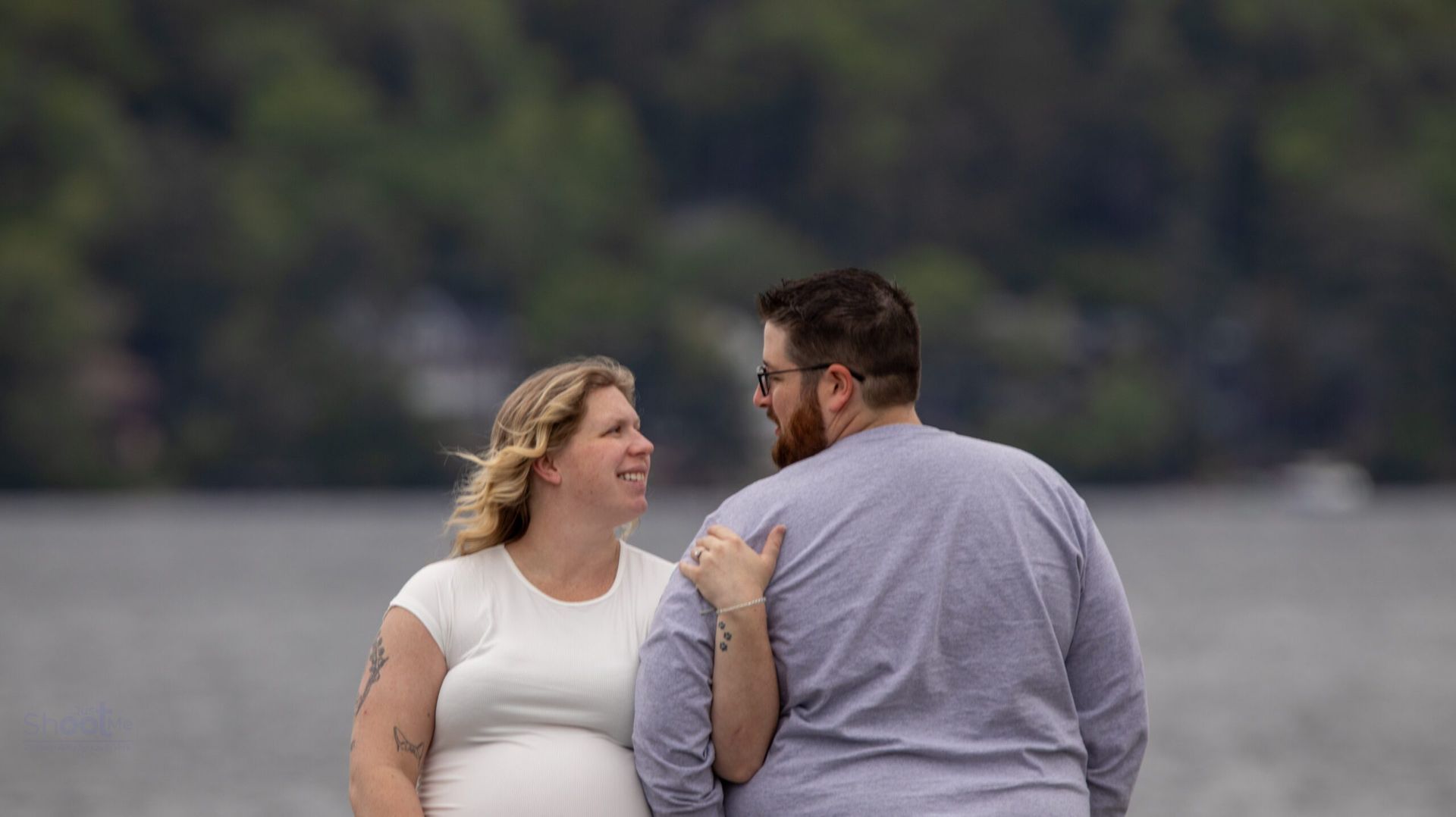 A couple stands together by a body of water, smiling at each other.