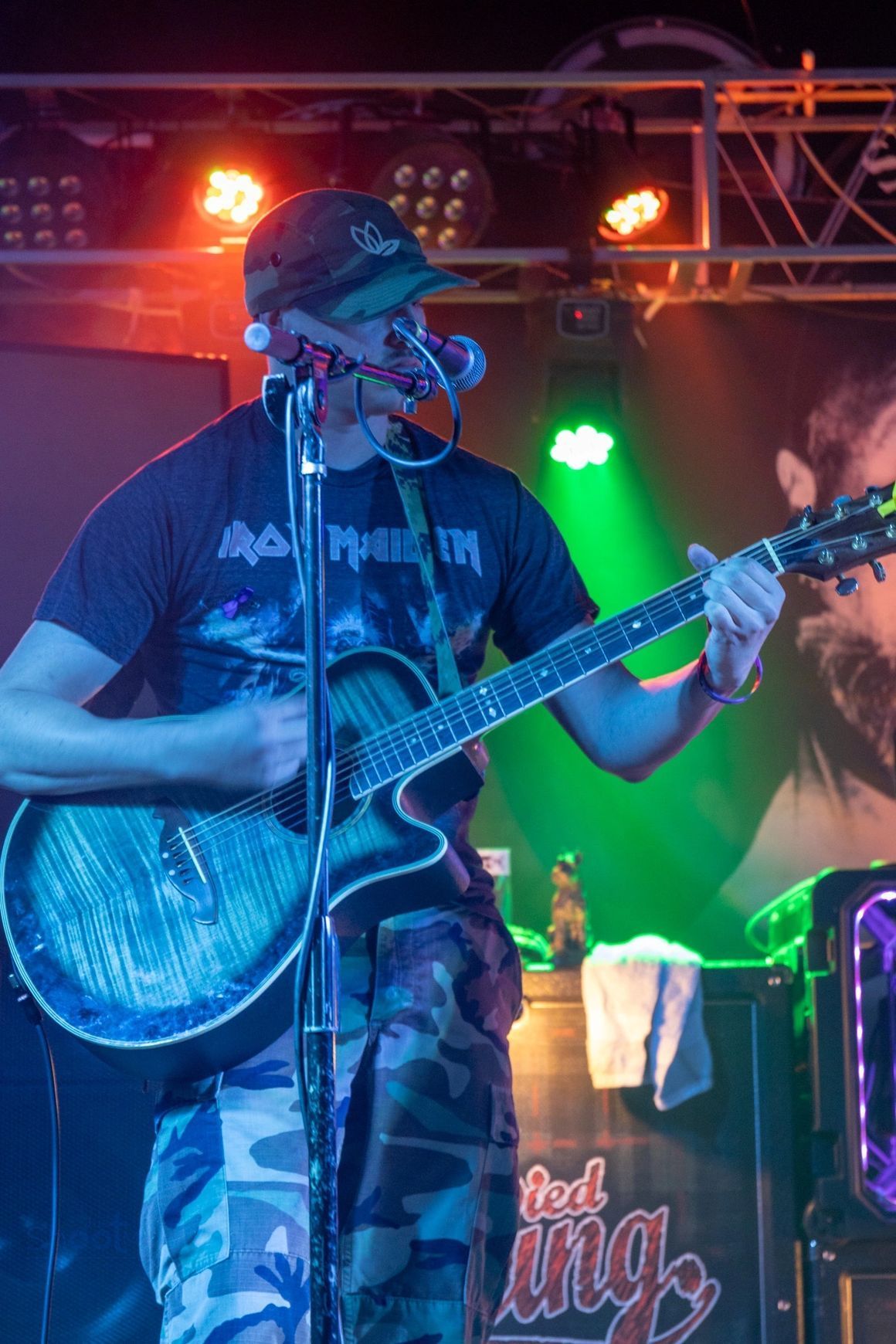 A musician in a cap and camo pants playing an acoustic guitar and singing into a microphone on a stage with stage lights.