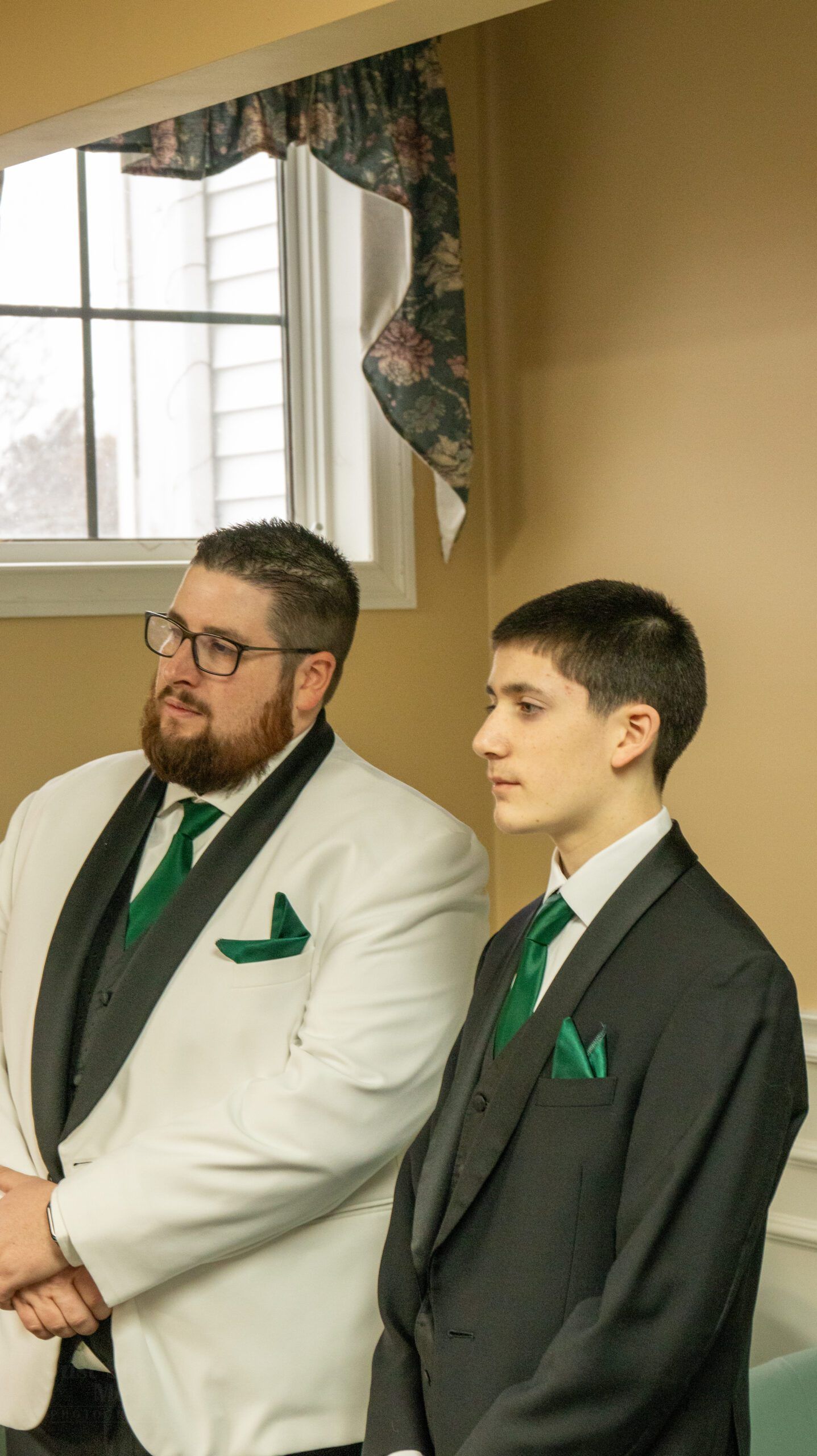 Two people wearing formal suits with green ties and pocket squares stand side-by-side near a window with a floral valance.