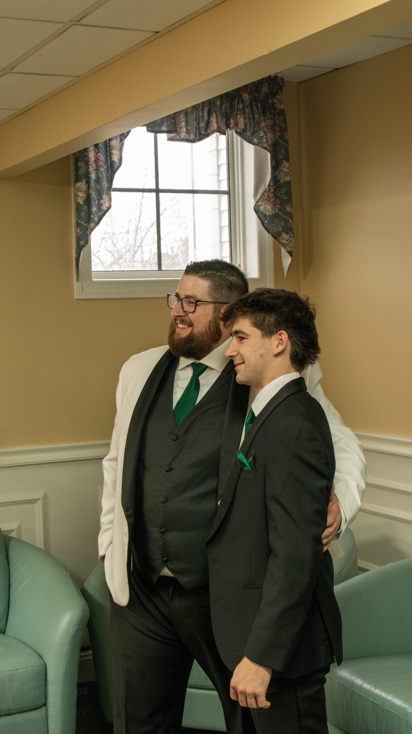 Two men in suits and green ties pose together in an indoor room with light-colored chairs and a window with a valance.