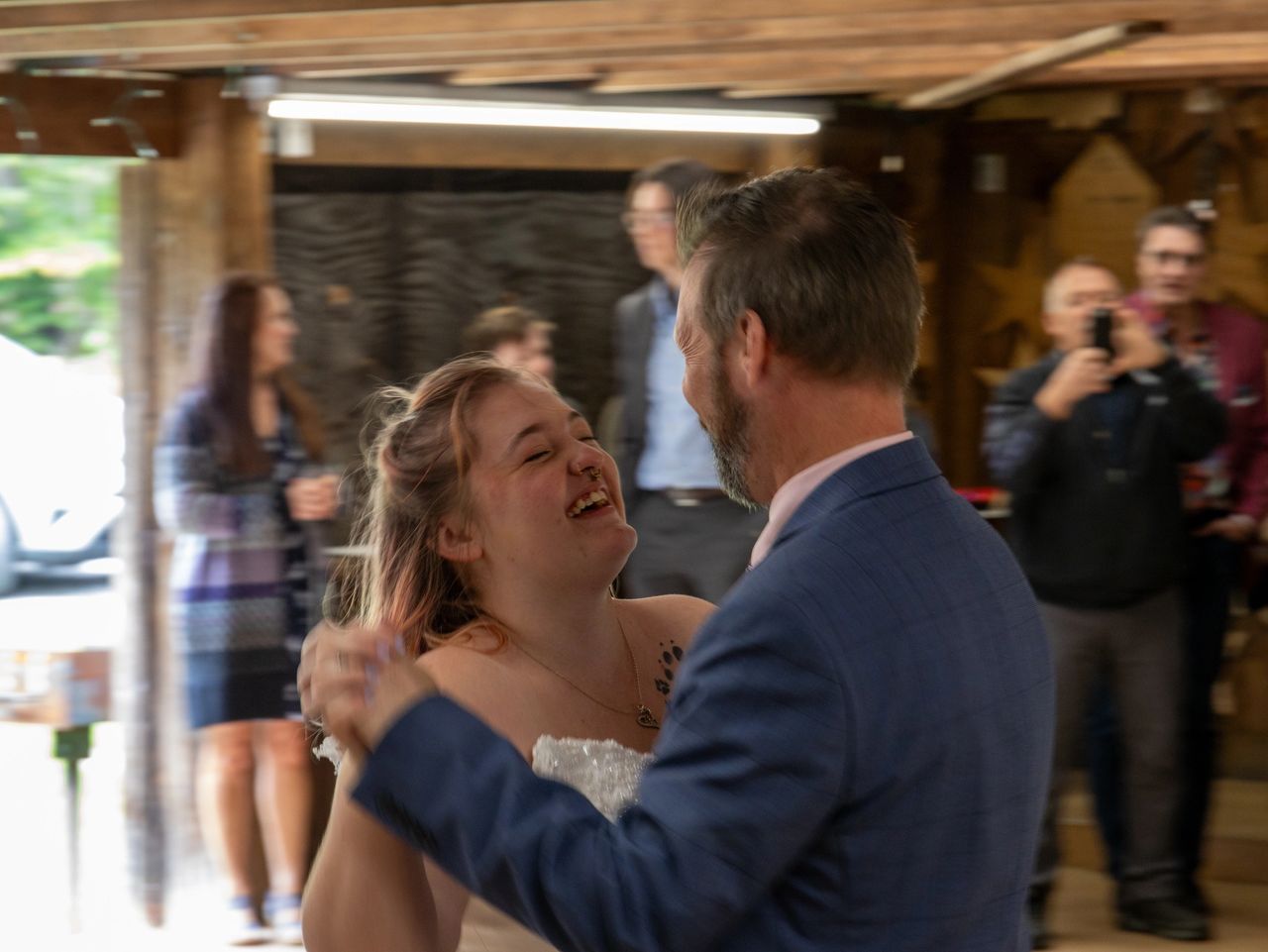 A bride laughing while dancing with a person in a blue suit inside a wooden structure, with onlookers in the background.