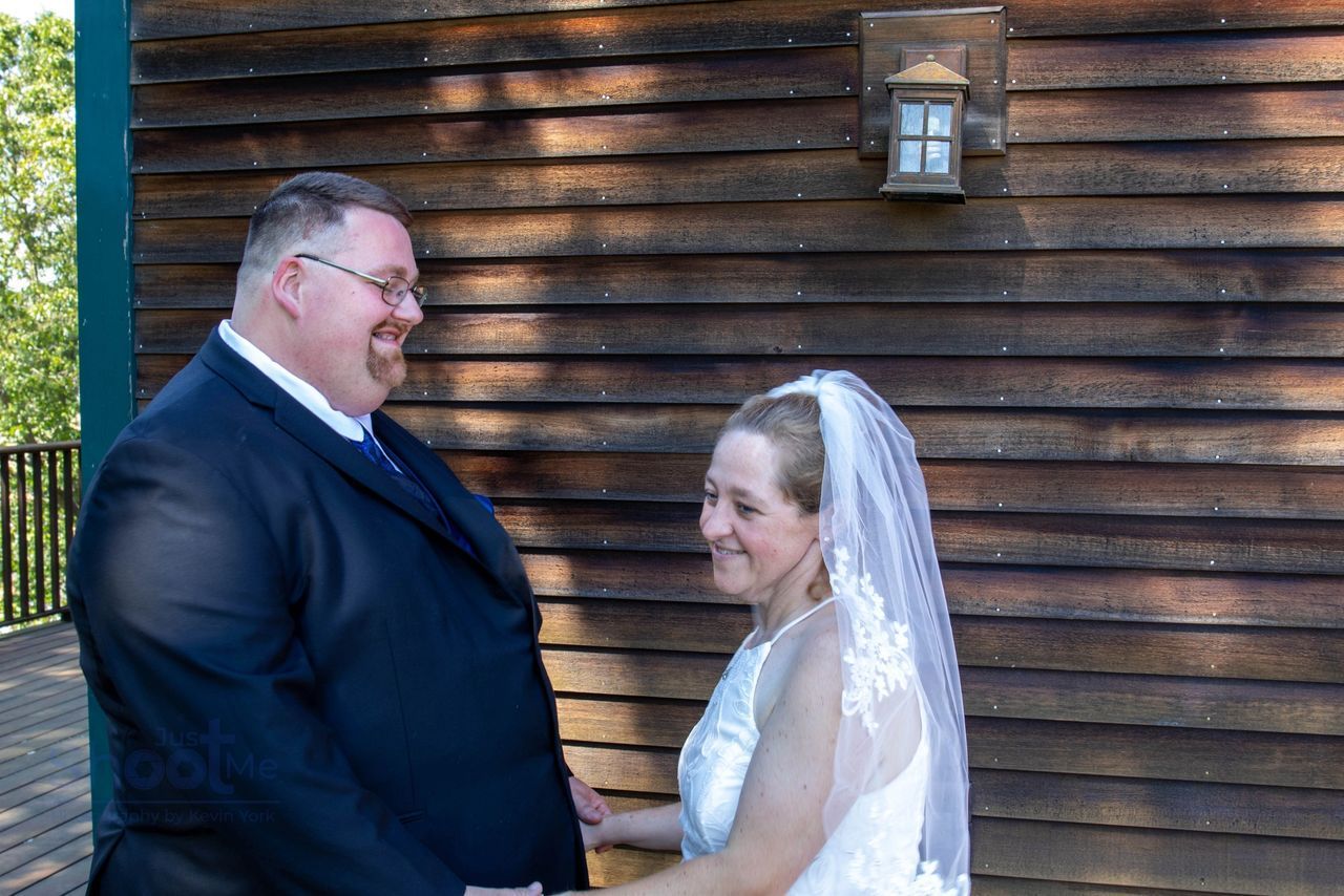 A couple in wedding attire smiles at each other while standing on a deck in front of a wood-sided building.