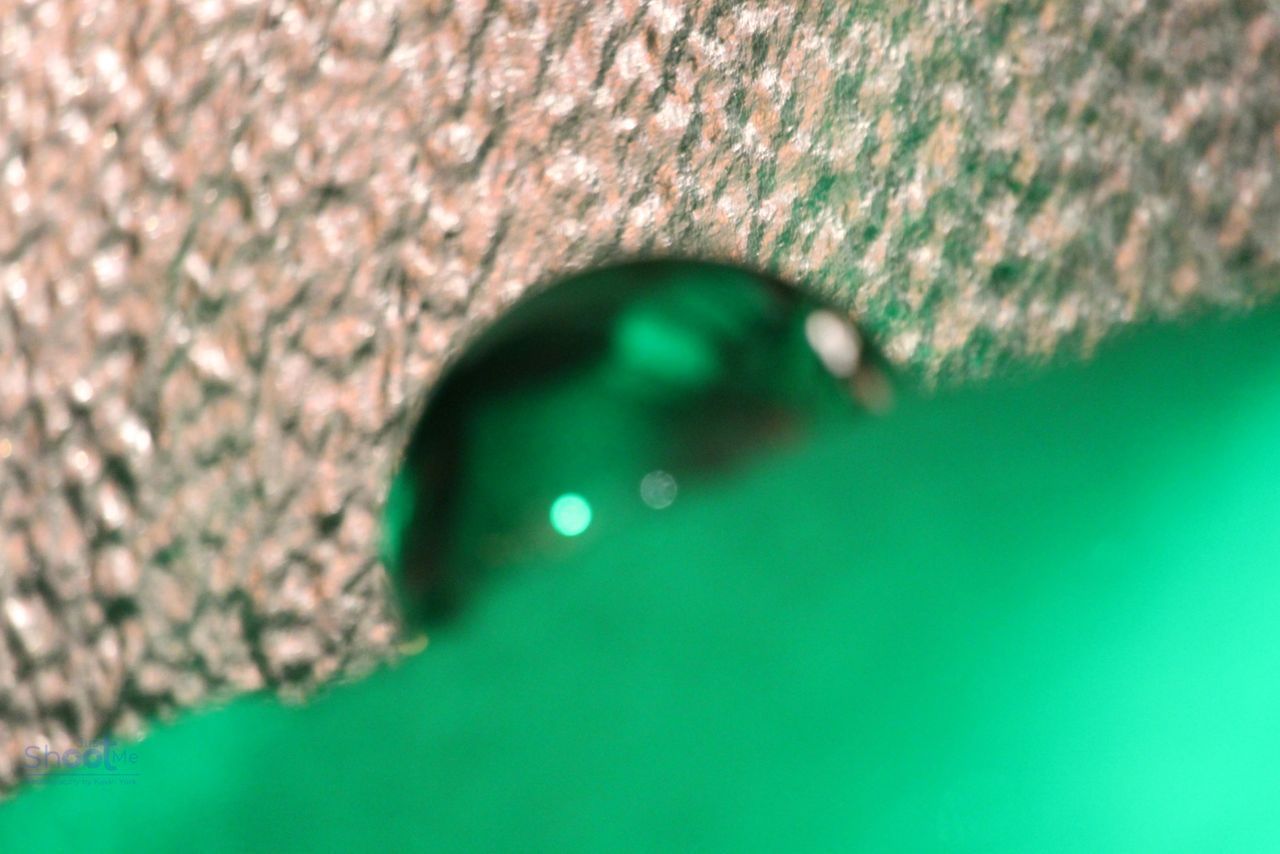 A clear water droplet clings to the edge of a textured, silver-colored surface against a bright, solid green background.