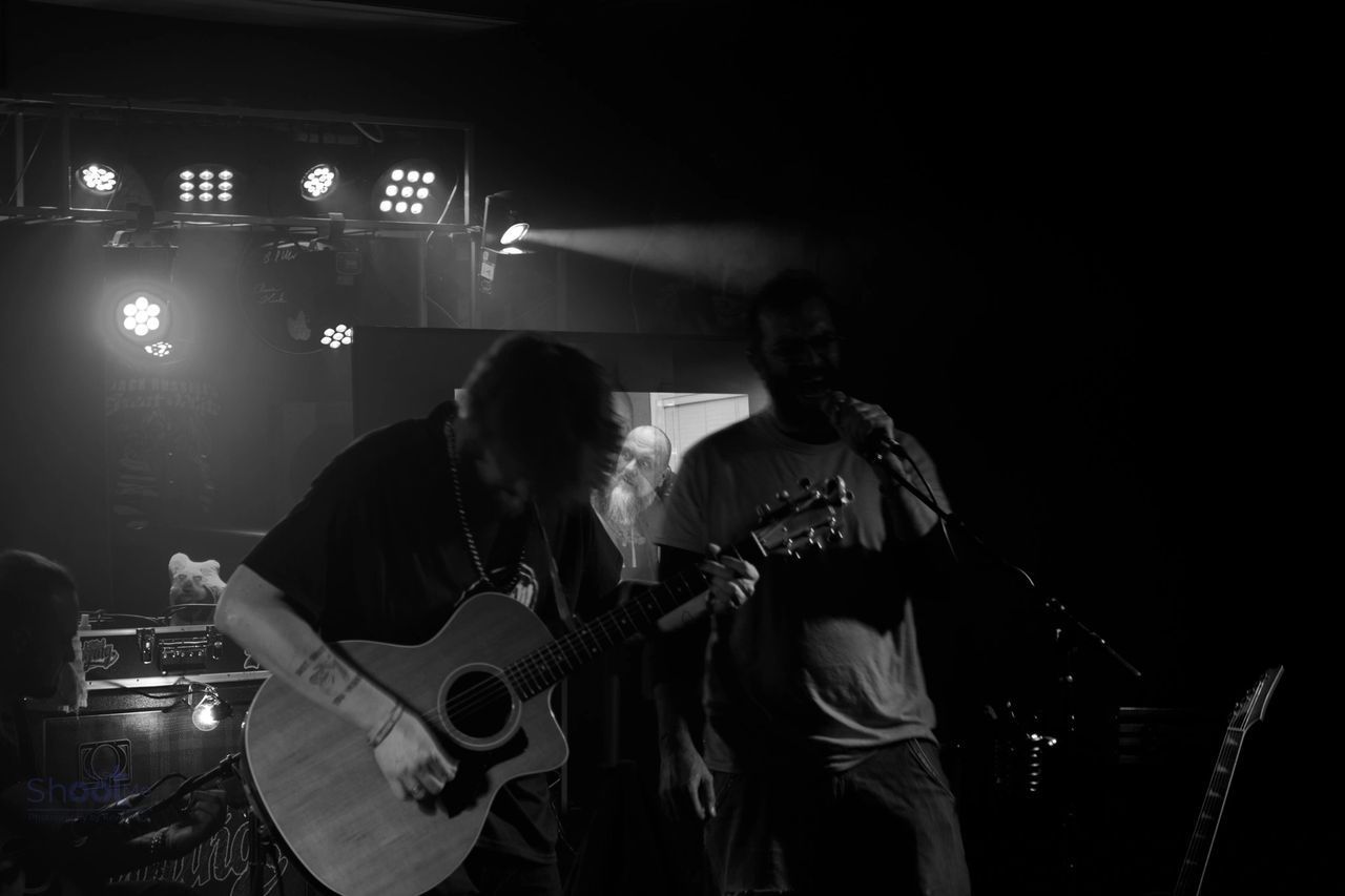 Two musicians performing on a dark, dimly lit stage with spotlight beams; one plays an acoustic guitar, the other sings.