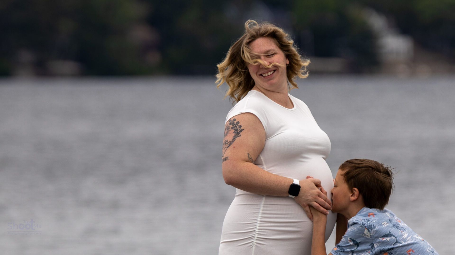 A person with a baby bump smiling while a child leans in to kiss their stomach near a body of water.
