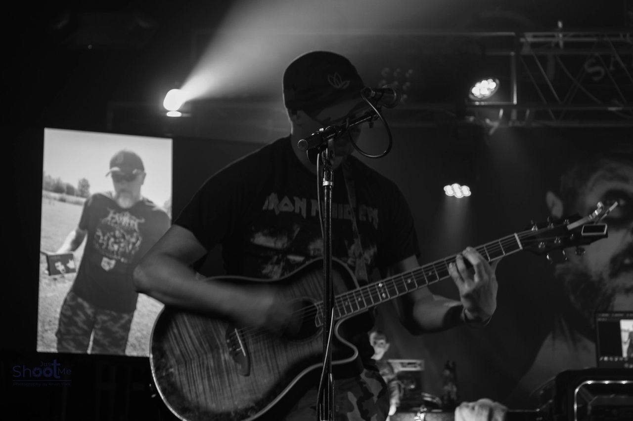 A musician plays an acoustic guitar on stage, with a projected image of a person behind them in a dark, lit venue.