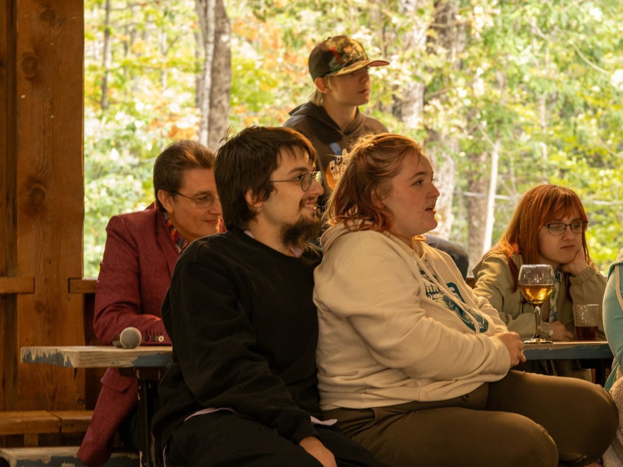 A group of people sit at tables in an outdoor, wooded setting, focused on an event with glasses and a microphone nearby.
