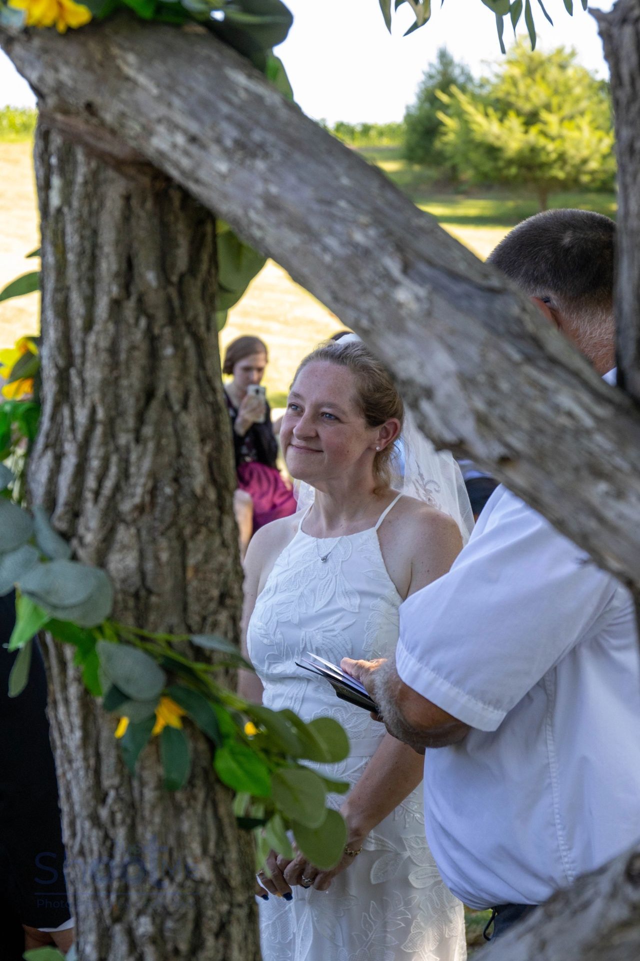 A person in a white dress stands outdoors during a ceremony, viewed through a tree frame as they look toward an officiant.