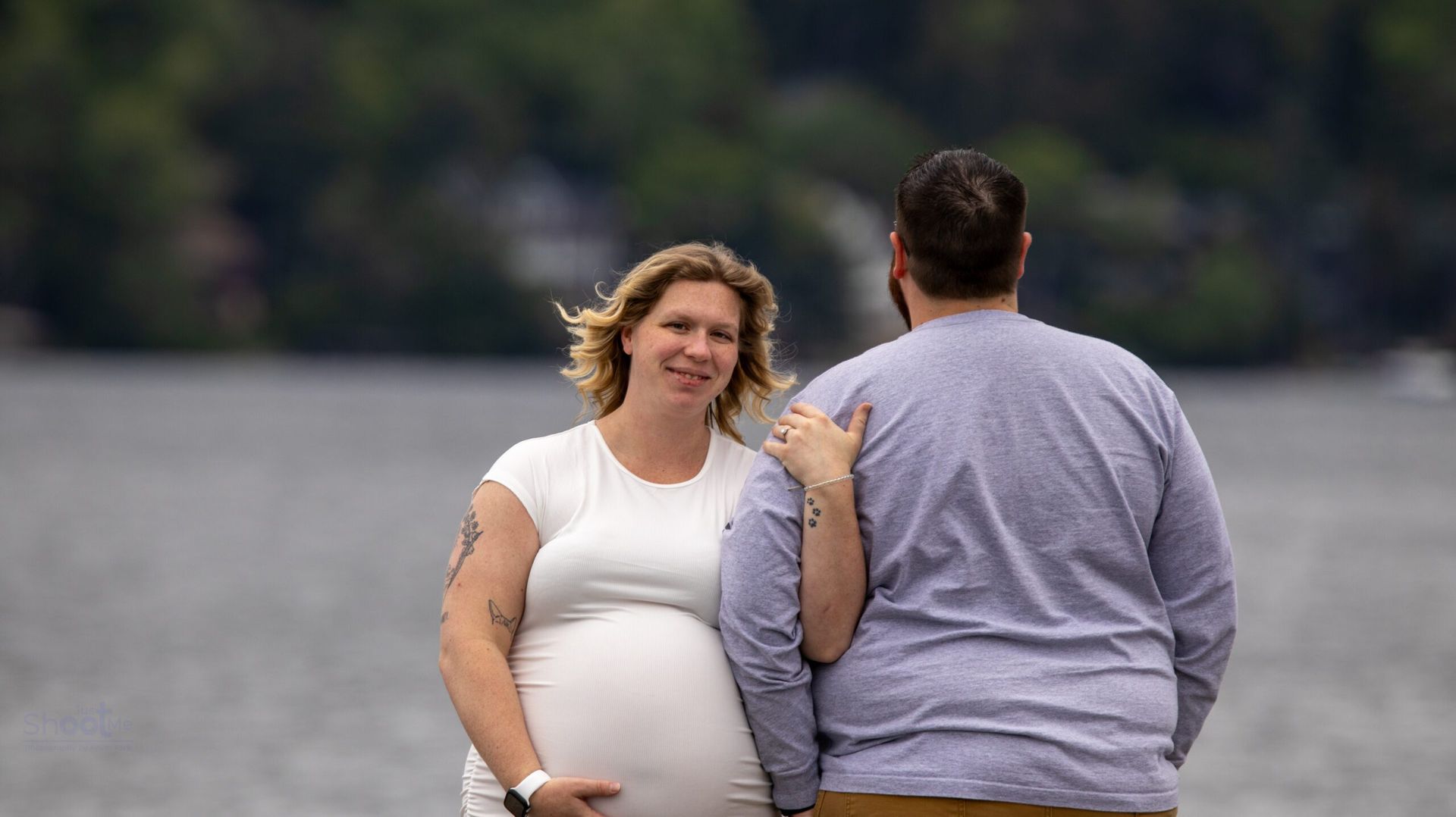 A pregnant woman and her partner stand together near a lake, looking toward the camera.