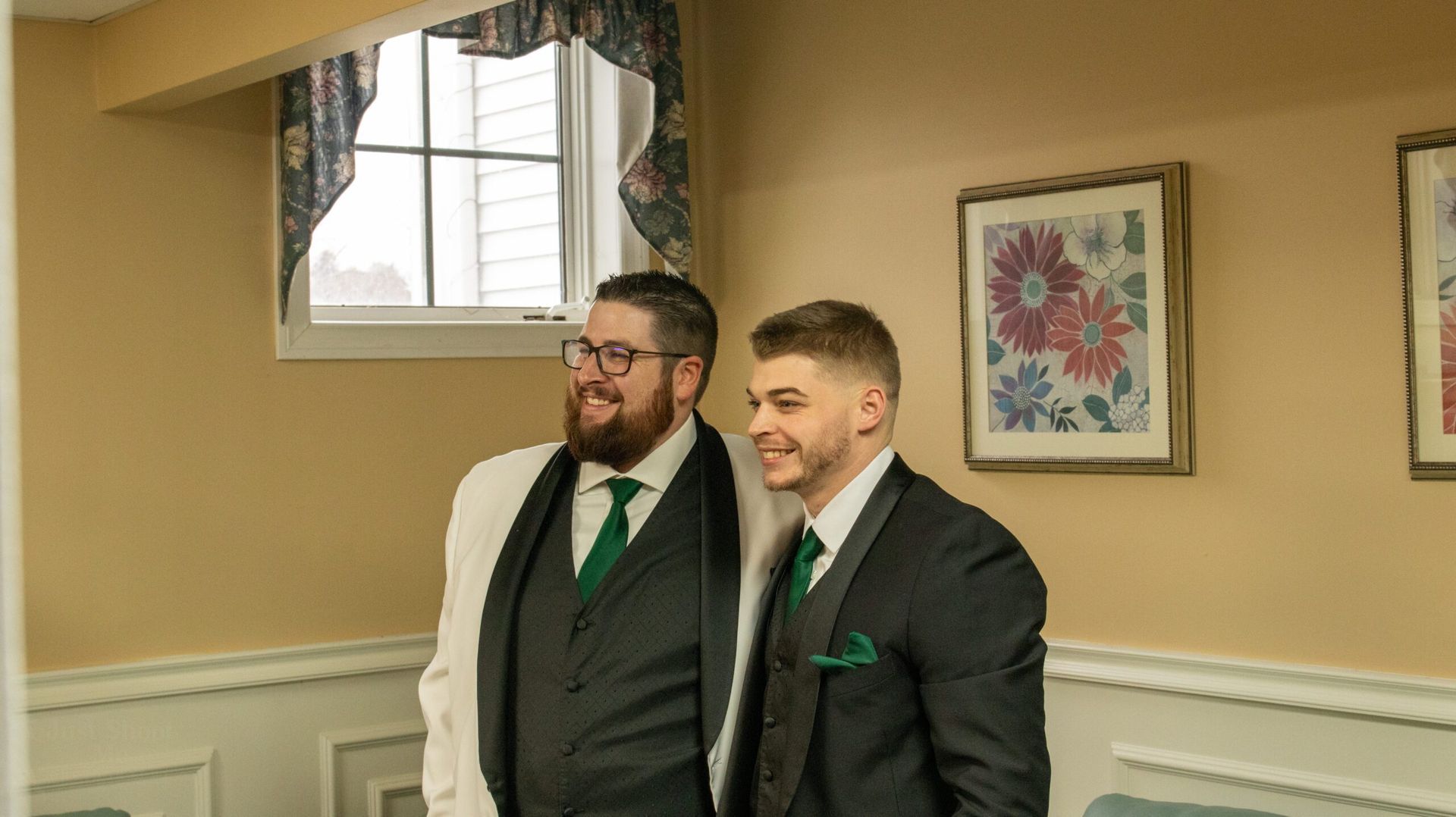 Two men in dark suits and green ties smile while standing together in a room with tan walls and floral wall art.