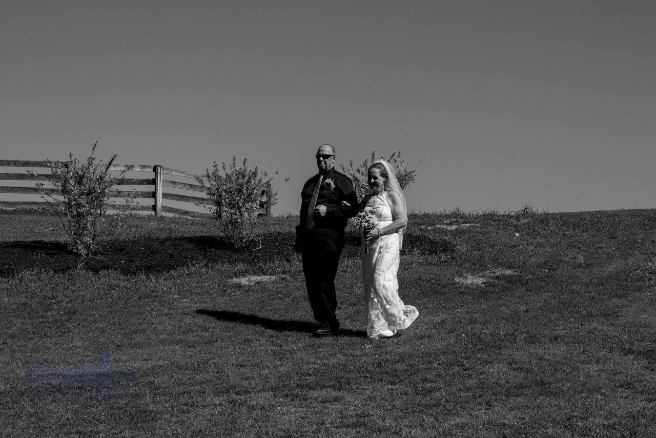 A person in a dark suit and a person in a white wedding gown walk together across a grassy field near a wooden fence.