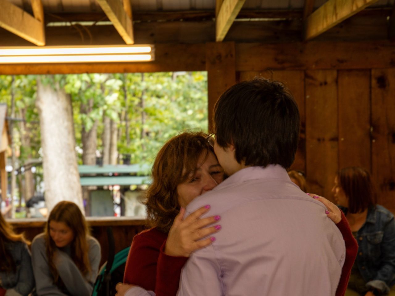 A person in a maroon sweater hugs another individual in a lavender button-down shirt in an indoor, wood-paneled space.