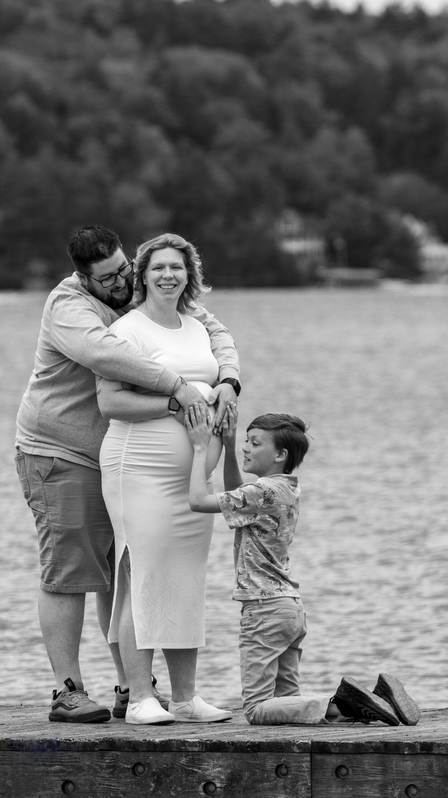 A family stands on a dock by a lake, hugging and holding a pregnant person's belly, set against a tree-lined background.
